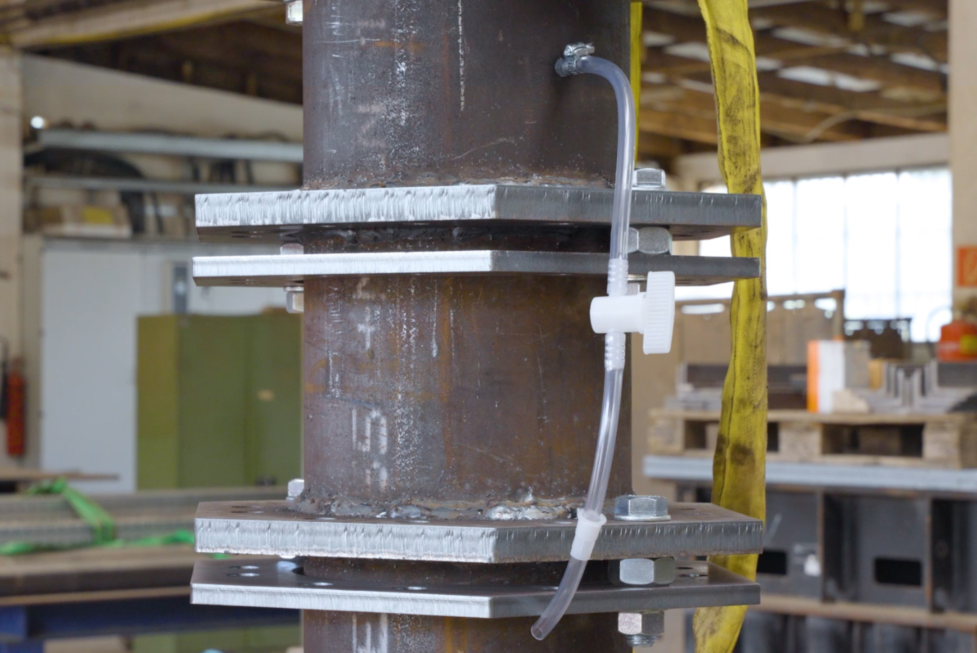Detalhe de coluna metálica cinzenta com fitas brancas de marcação, tubagens azuis e brancas presas com grampos, em ambiente industrial com estruturas verdes e amarelas ao fundo