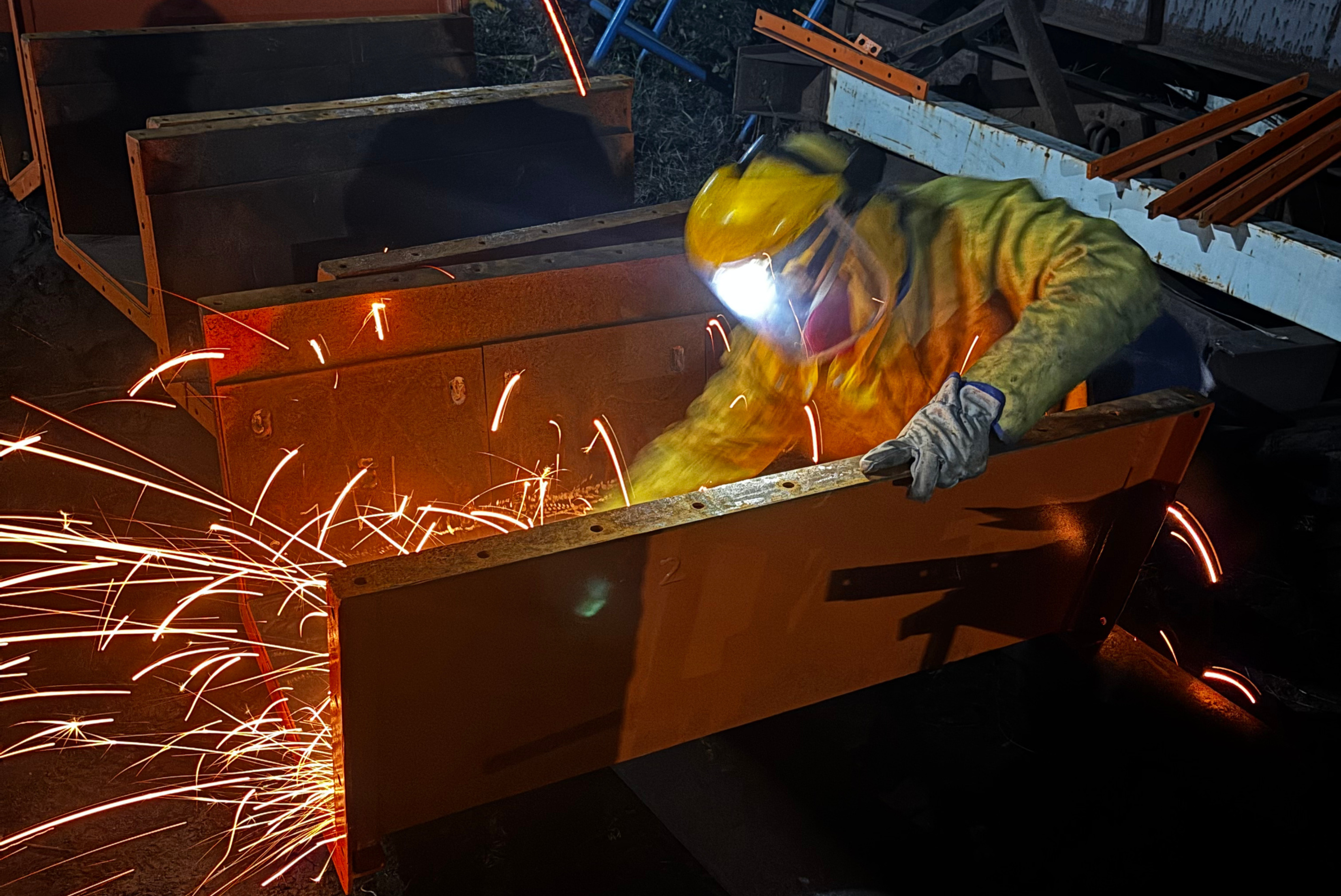 Trabalhador em equipamento de proteção amarelo e capacete realizando soldagem em estrutura metálica de laranja, com sparks de solda voando em torno da peça de trabalho