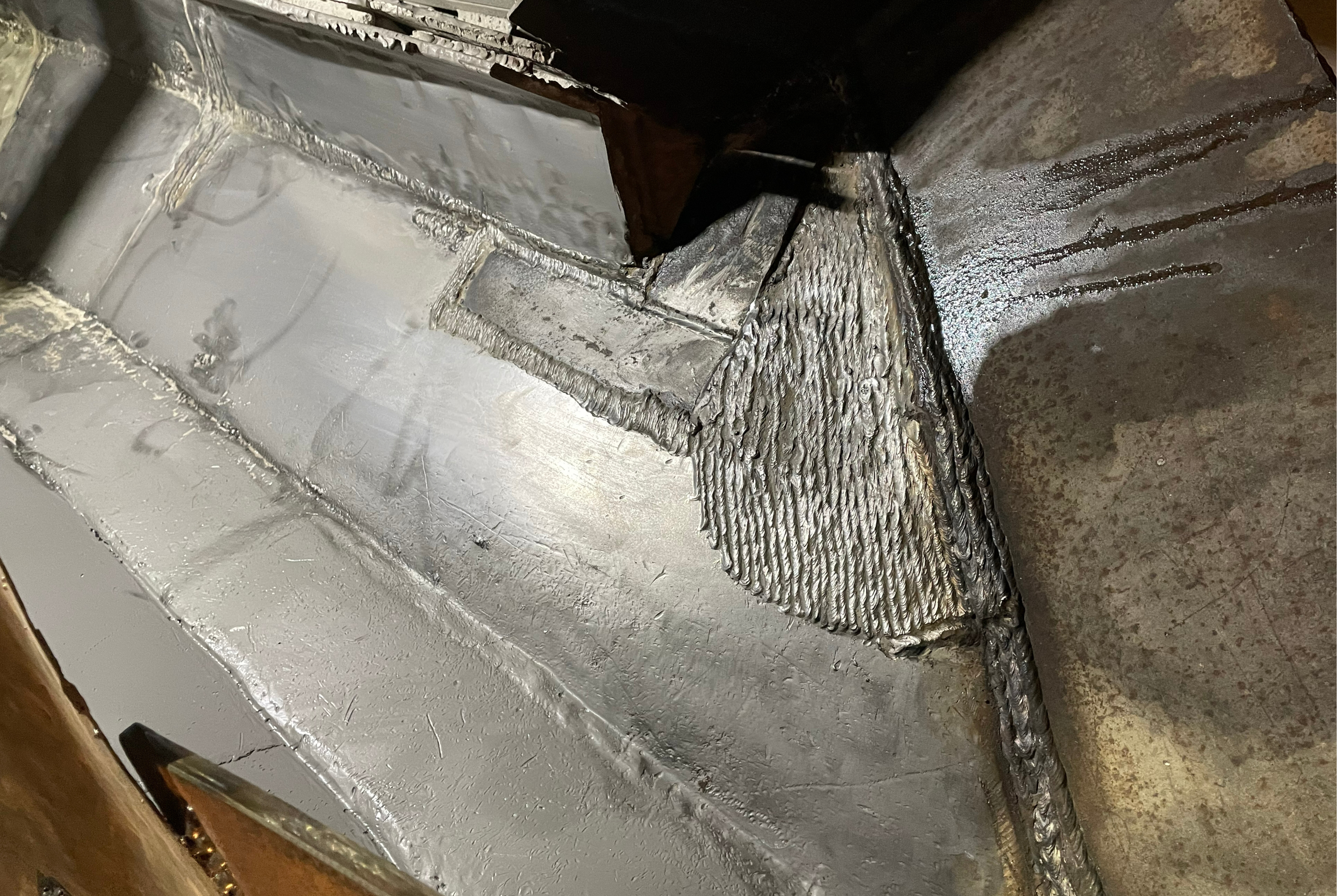 Detalhe de costura soldada em componente industrial metálico cinzento com revestimento protetor, mostrando padrão de soldadura texturizado e acabamento em várias camadas de material.
