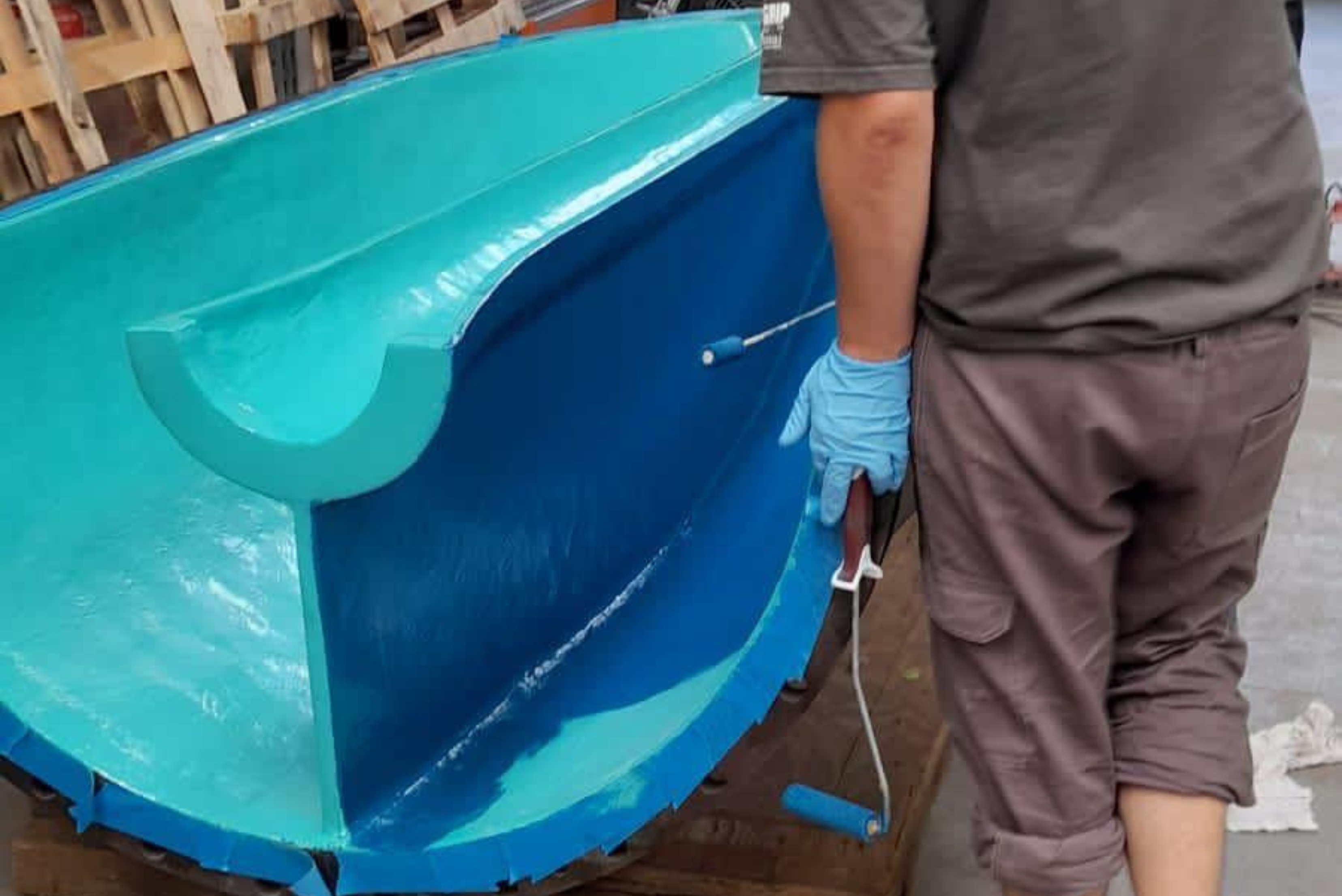 Trabalhador enluvado em azul inspecionando ou aplicando acabamento em componente industrial azul claro em forma de meia-tigela, com superfícies lisas e revestidas em ambiente de fábrica."