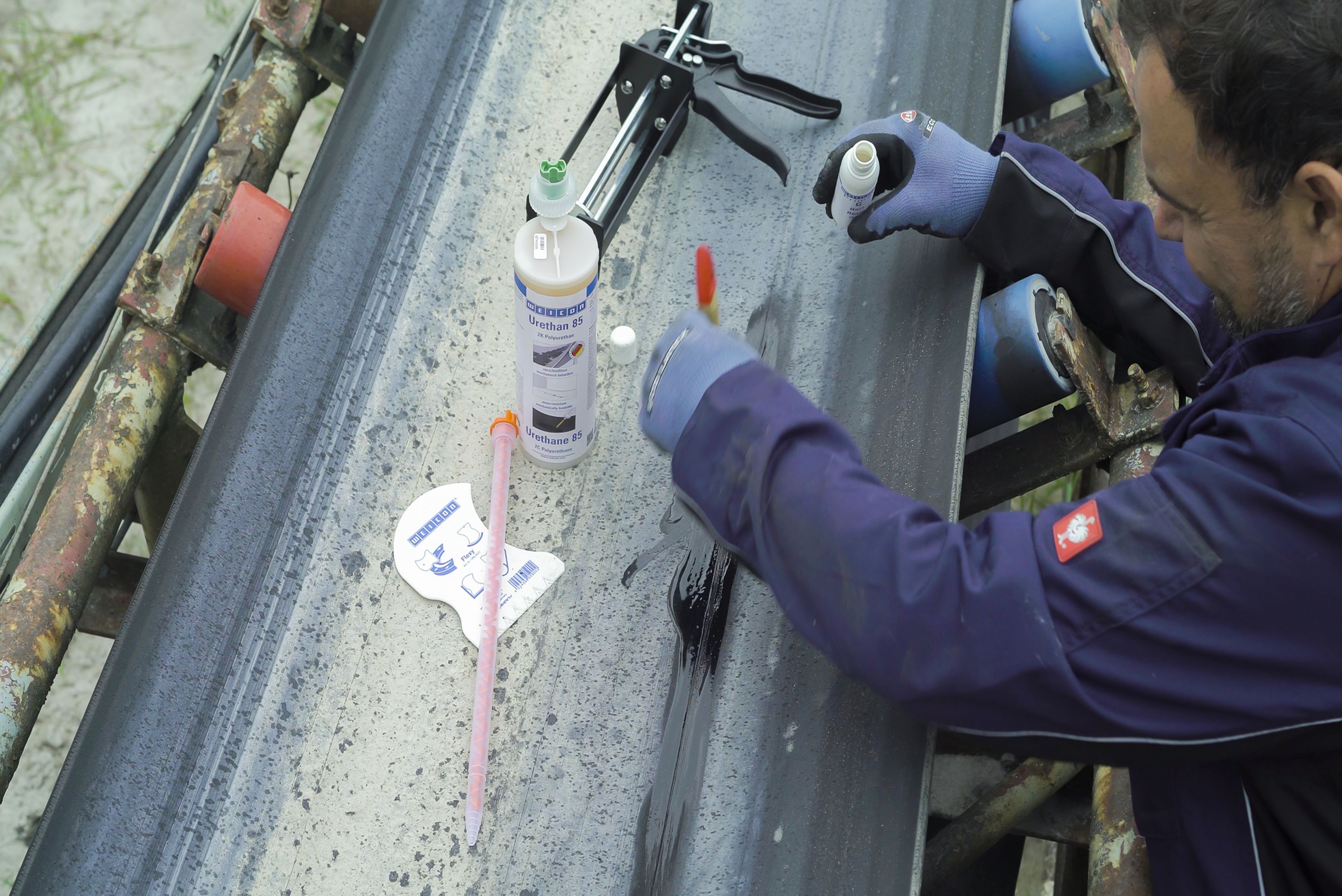 Trabalhador em uniforme azul realizando medição ou marcação em superfície metálica cinzenta com ferramenta de medida, com frascos de produto e materiais dispostos no local de trabalho