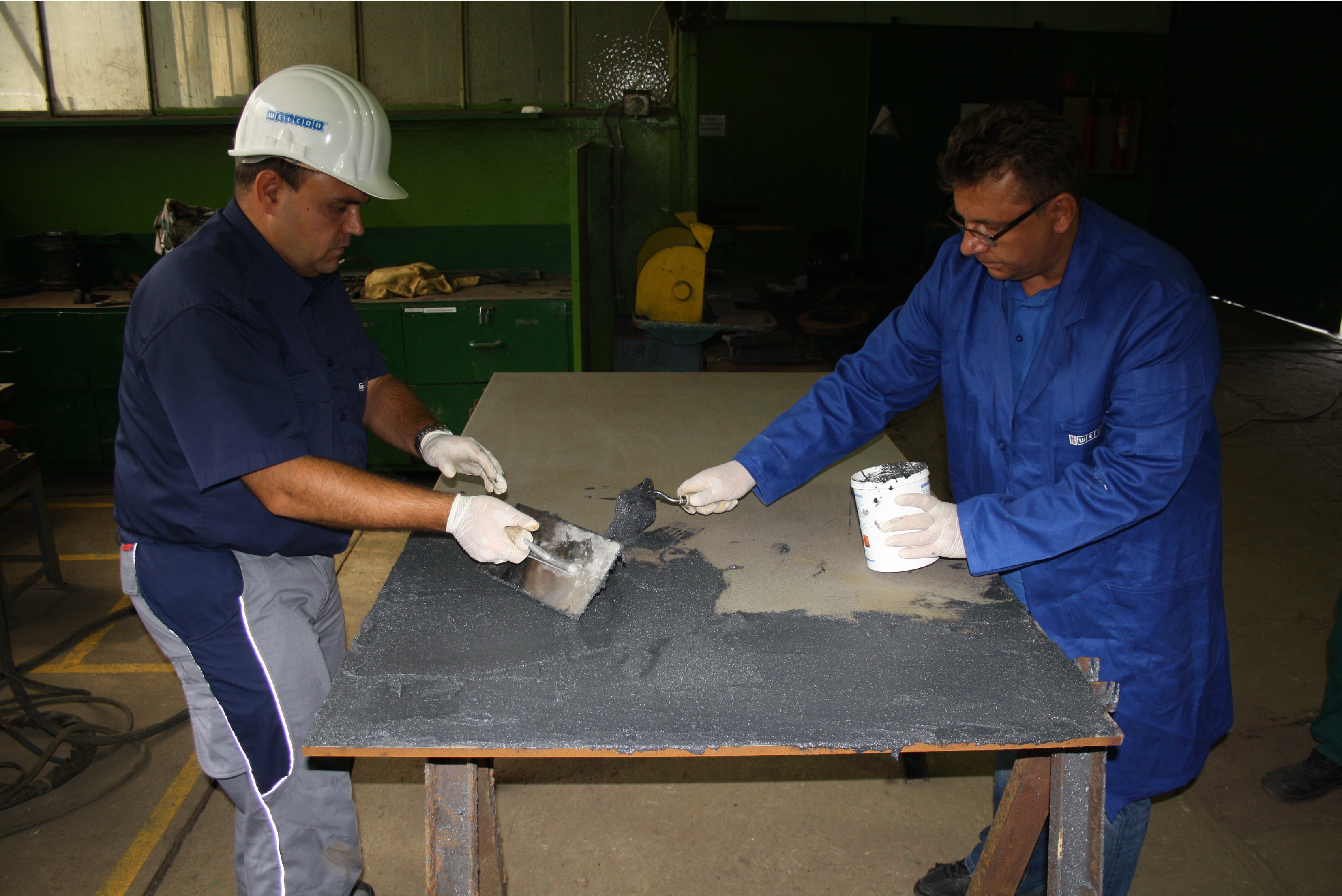 Dois trabalhadores em uniformes azuis e branco examinando e manuseando uma peça de metal cinzento sobre uma mesa de trabalho em ambiente industrial com estruturas verdes ao fundo.