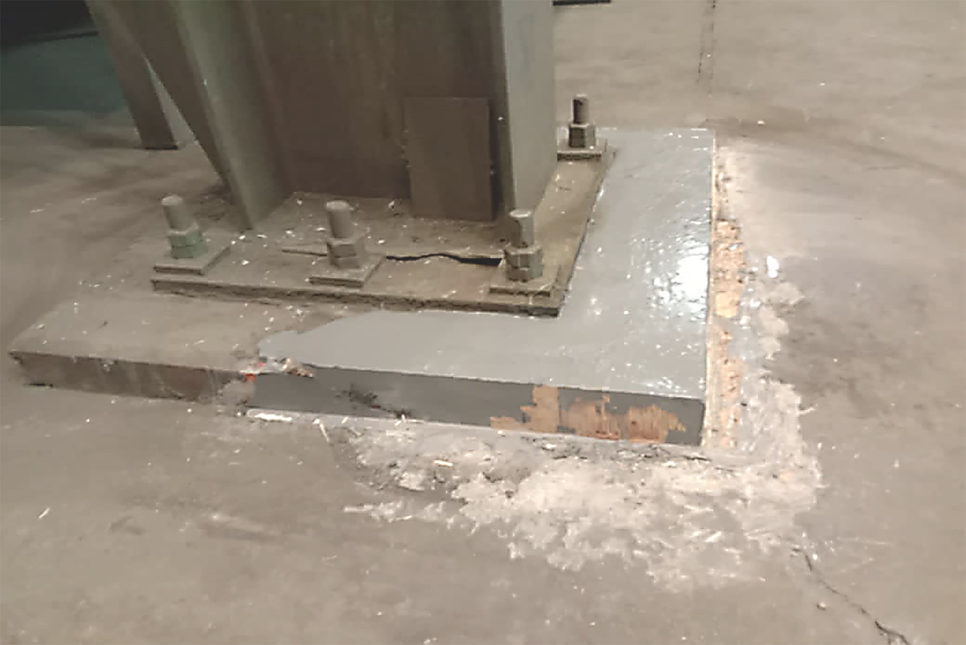 Uma base de pilar de aço industrial robusta, fixada com grandes parafusos e porcas a um bloco de fundação de concreto cinza, localizado em um piso de fábrica, com áreas de desgaste e reparo visíveis ao redor da base.