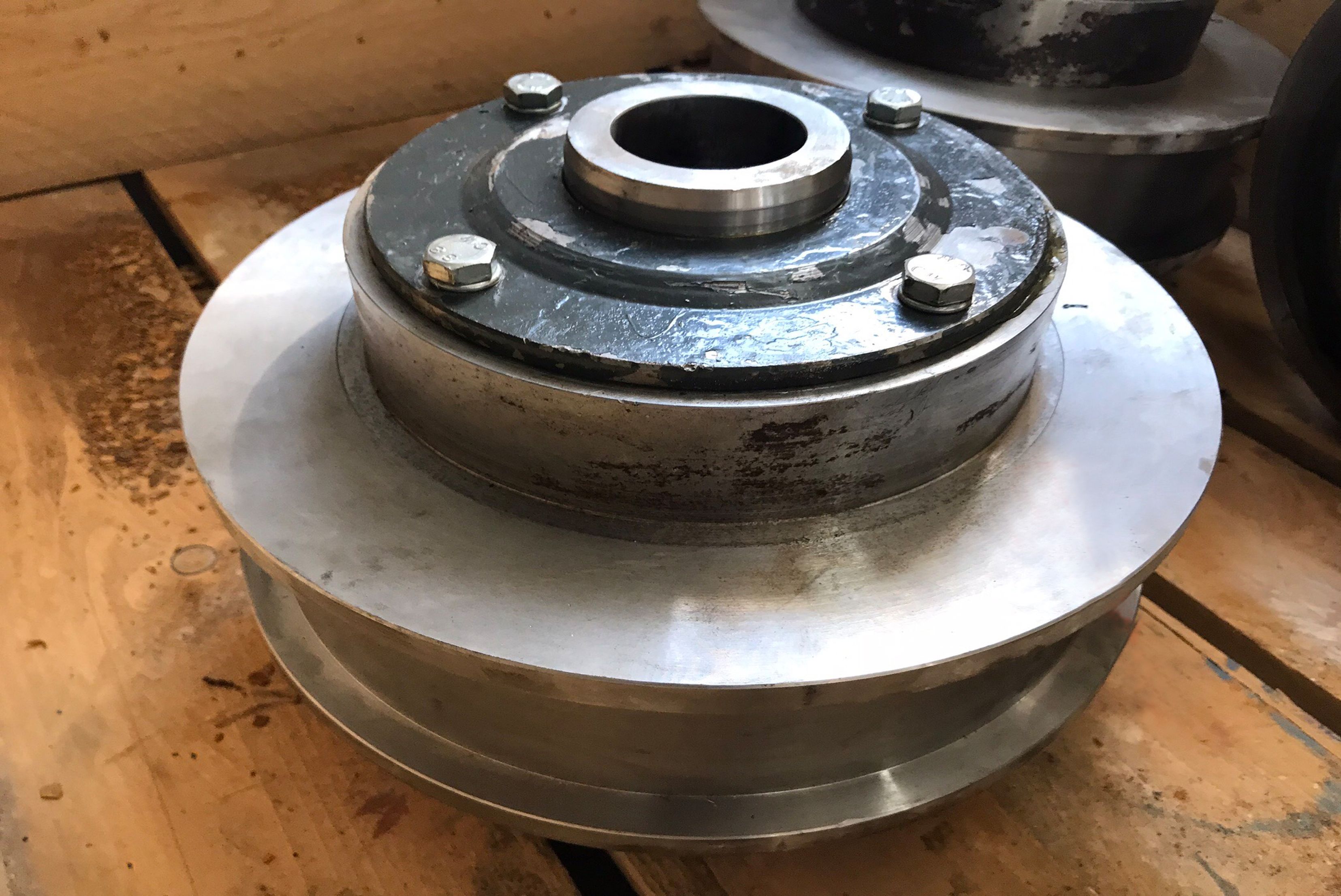 Componente industrial em forma de disco com múltiplas camadas metálicas, eixo central com furos de fixação, núcleo superior cinzento escuro e base branca brilhante, apoiado em superfície de madeira