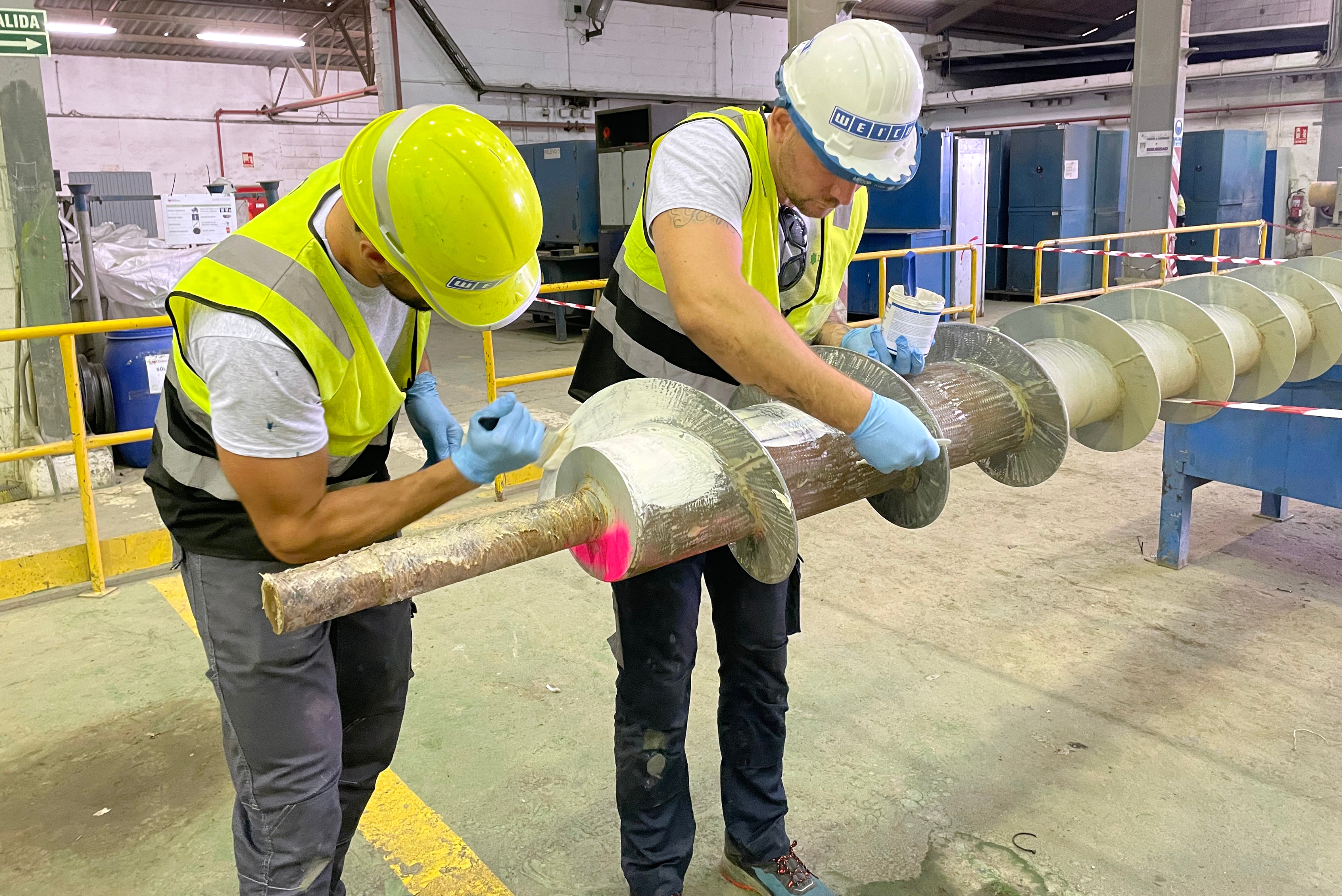 Dois trabalhadores em uniformes amarelos fluorescentes e capacetes brancos inspecionando cilindro industrial metálico com flanges, em ambiente de fábrica com demarcação amarela no piso e equipamentos ao fundo