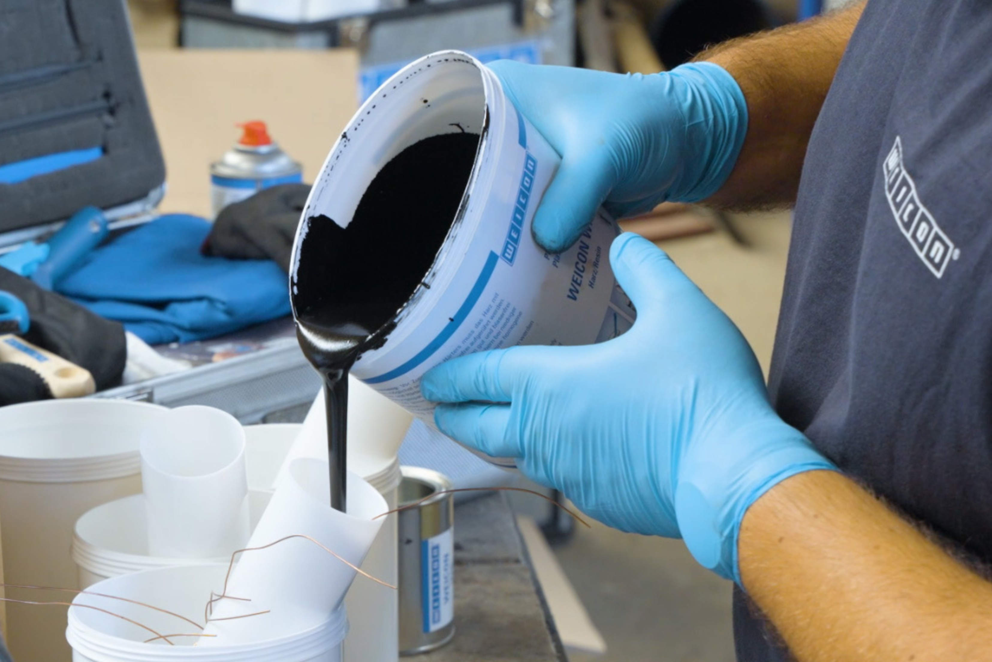 Mão enluvada em azul vertendo líquido escuro de um recipiente azul claro em um copo branco, em ambiente de laboratório ou industrial com equipamentos ao fundo