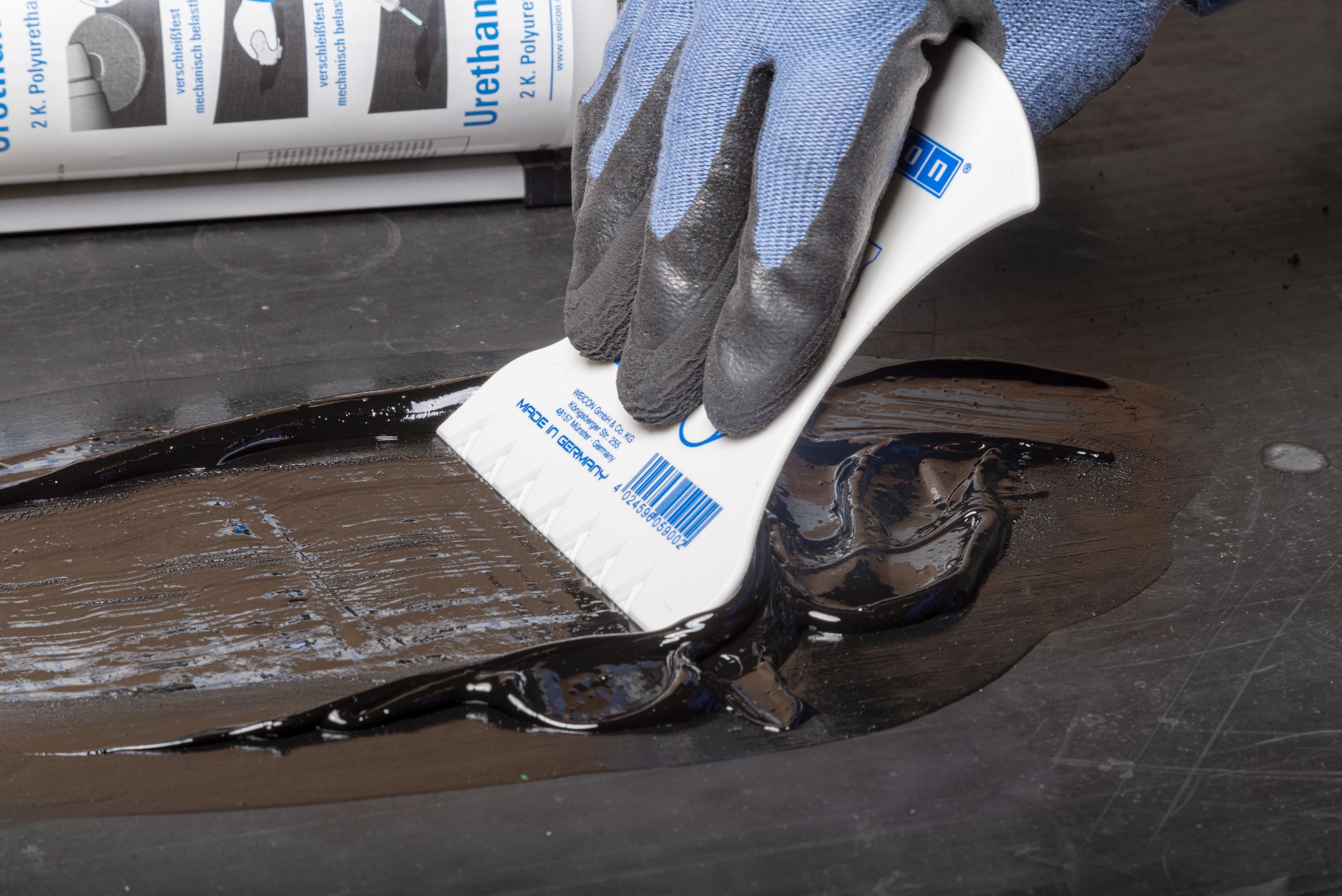 Mão enluvada em azul aplicando etiqueta ou rótulo branco com texto em superfície preta de componente industrial, com embalagem de produtos visível ao fundo