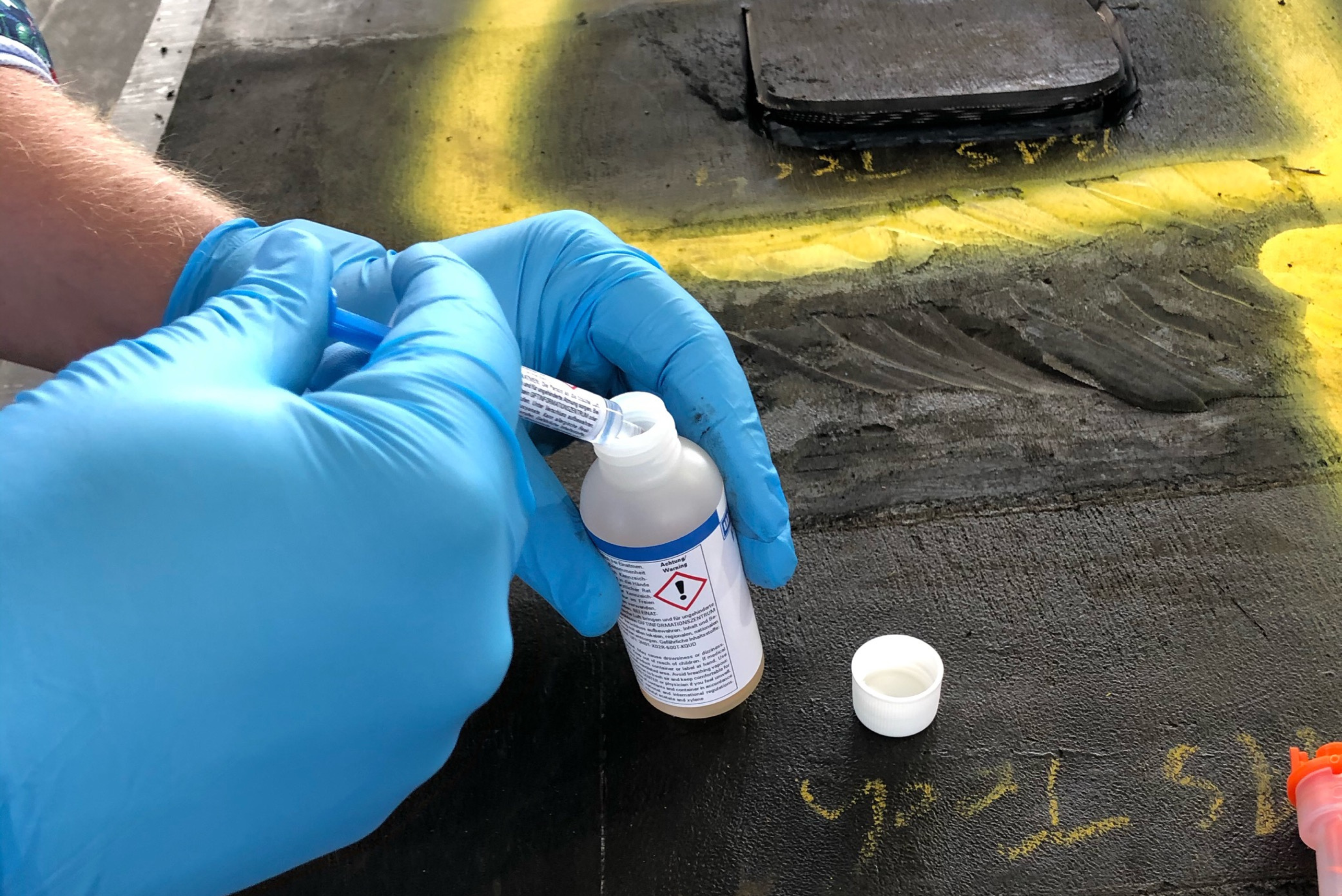 Uma pessoa com luvas azuis de proteção segura uma pequena garrafa plástica com um rótulo de advertência e usa uma seringa ou aplicador para adicionar líquido dentro dela, sobre uma superfície de trabalho escura com marcações amarelas.