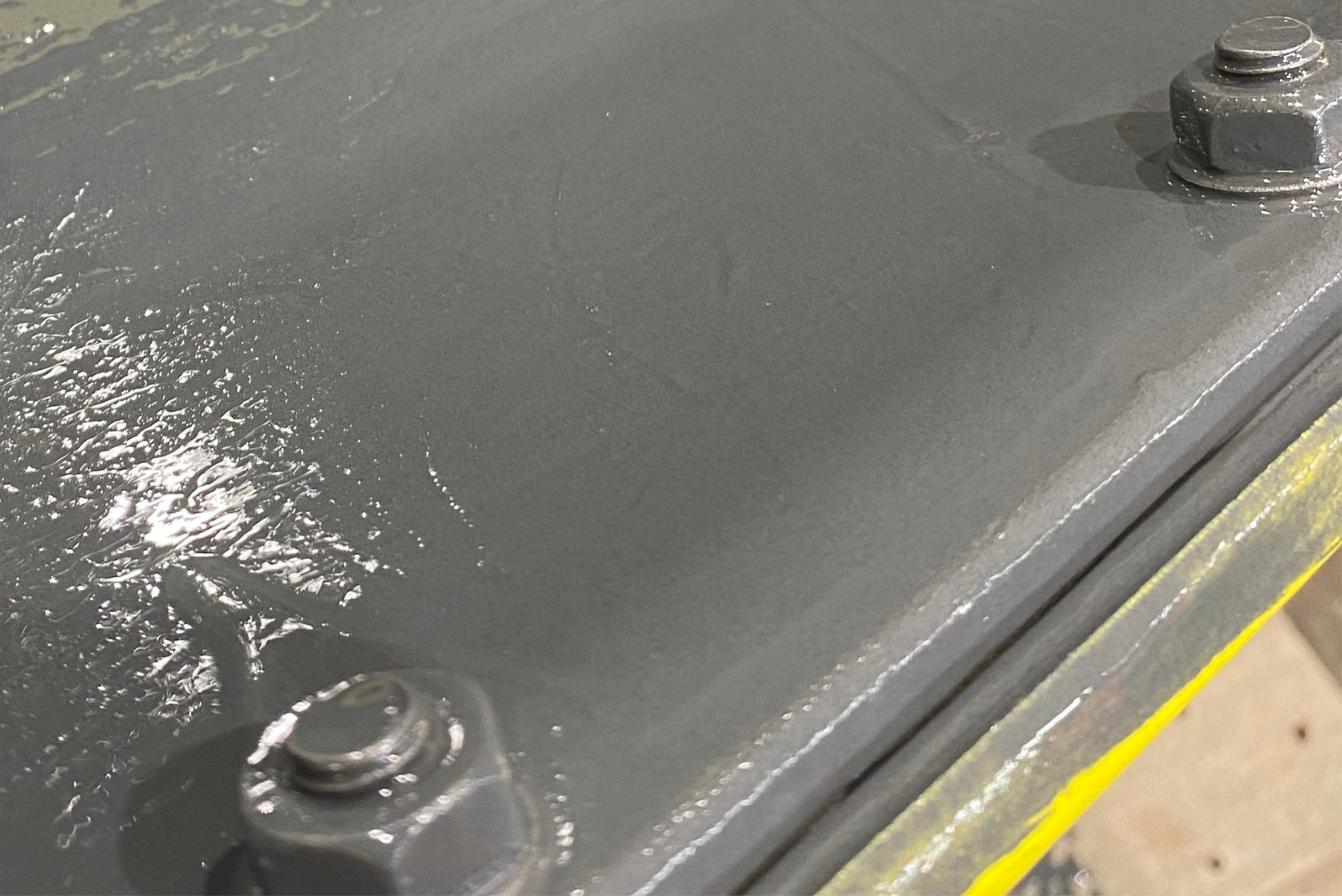 Superfície metálica cinzenta recém-revestida ou pintada com brilho úmido, mostrando parafusos de metal e fita adesiva amarela, em ambiente industrial.