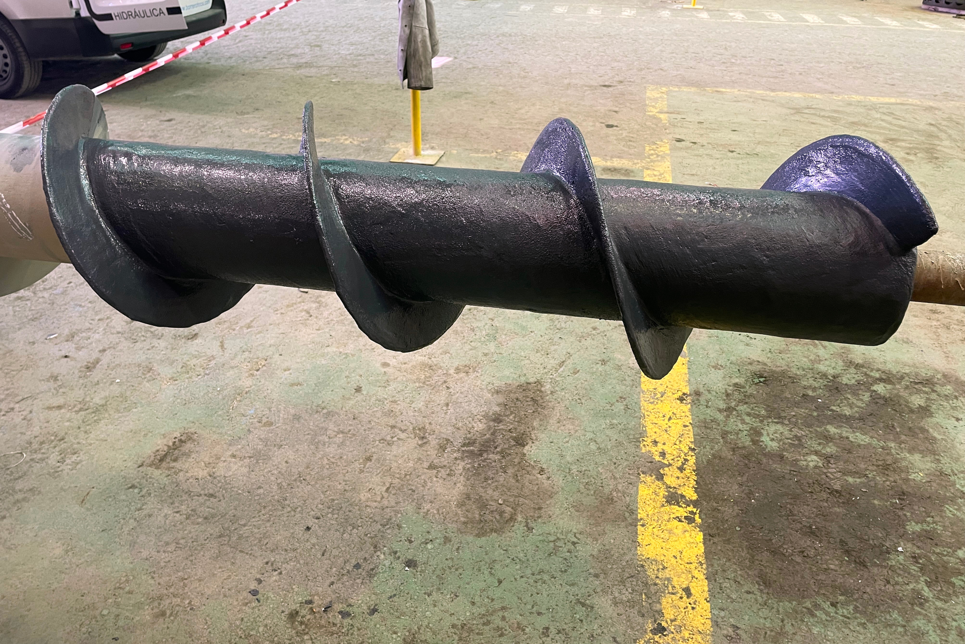 Cilindro industrial preto com seções segmentadas e discos de metal, deitado horizontalmente em piso cinzento com demarcação amarela, em ambiente de pátio ou área de armazenamento industrial.