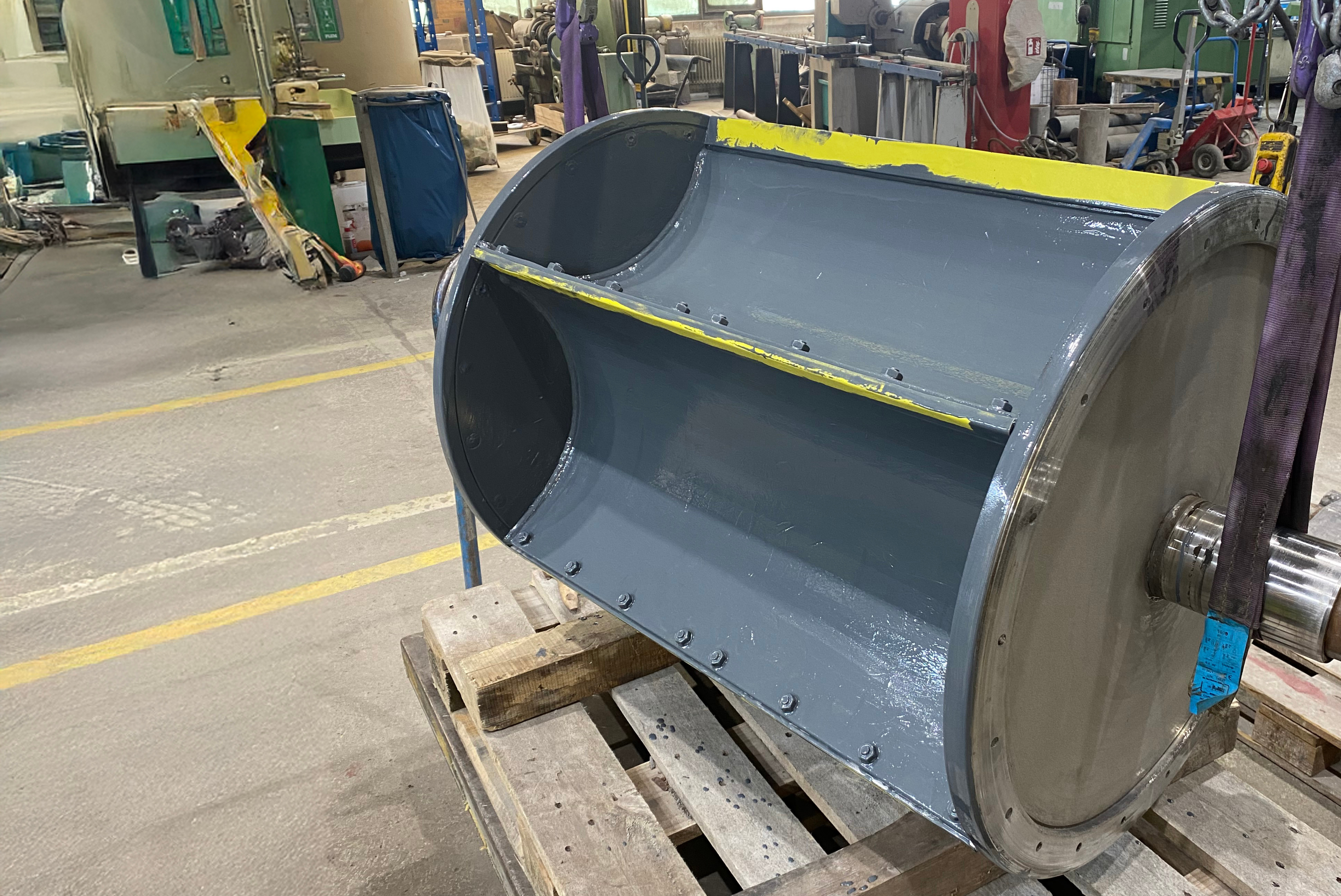 Cilindro industrial cinzento com borda superior amarela, aberturas circulares pretas laterais, montado em estrutura de transporte em pátio de fábrica com máquinas e equipamentos ao fundo.