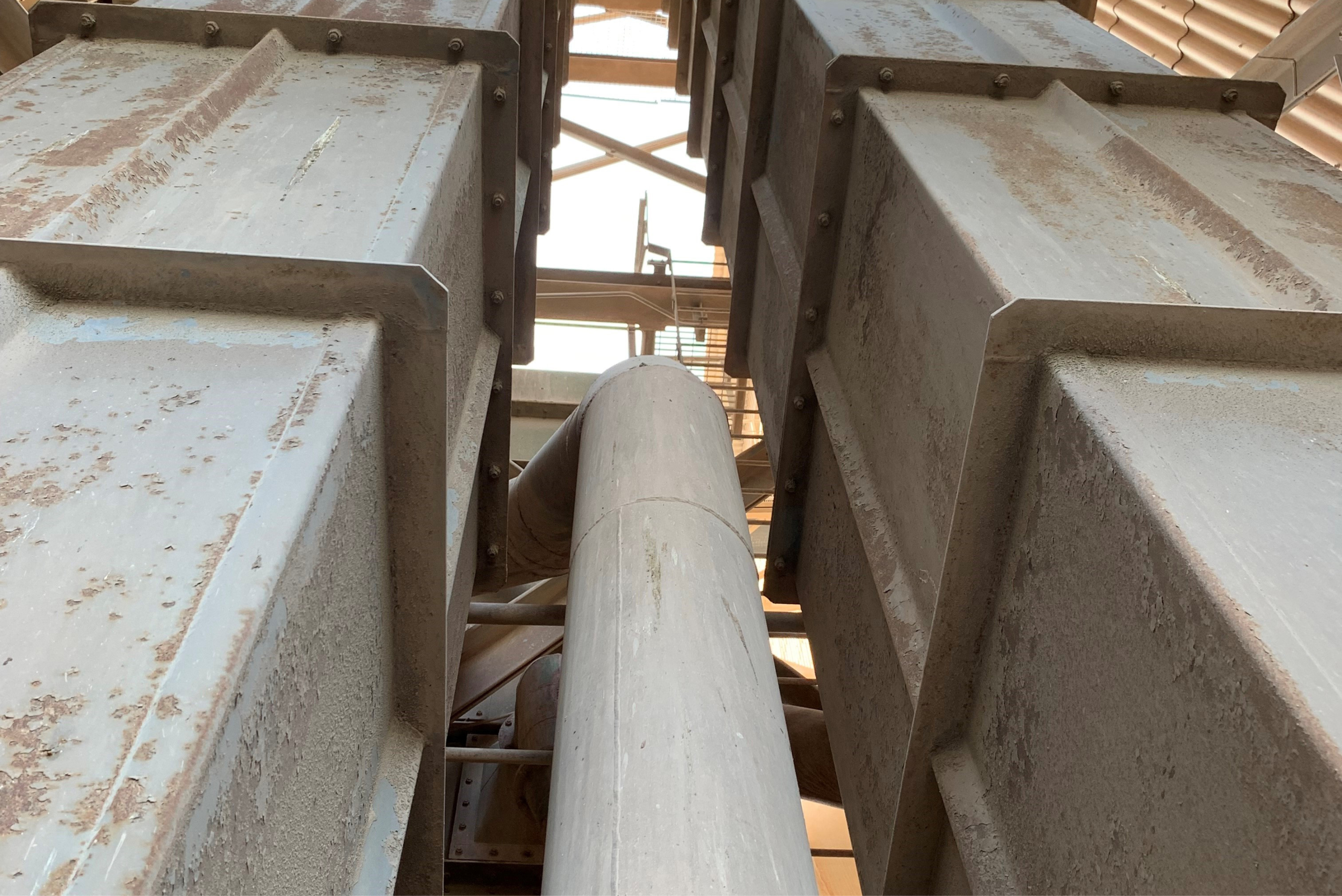 Vista longitudinal do interior de contêiner com paredes cinzentas de concreto ou metal, estrutura cilíndrica central, superfícies com marcas de ferrugem e desgaste, com aberturas de luz natural ao fundo.