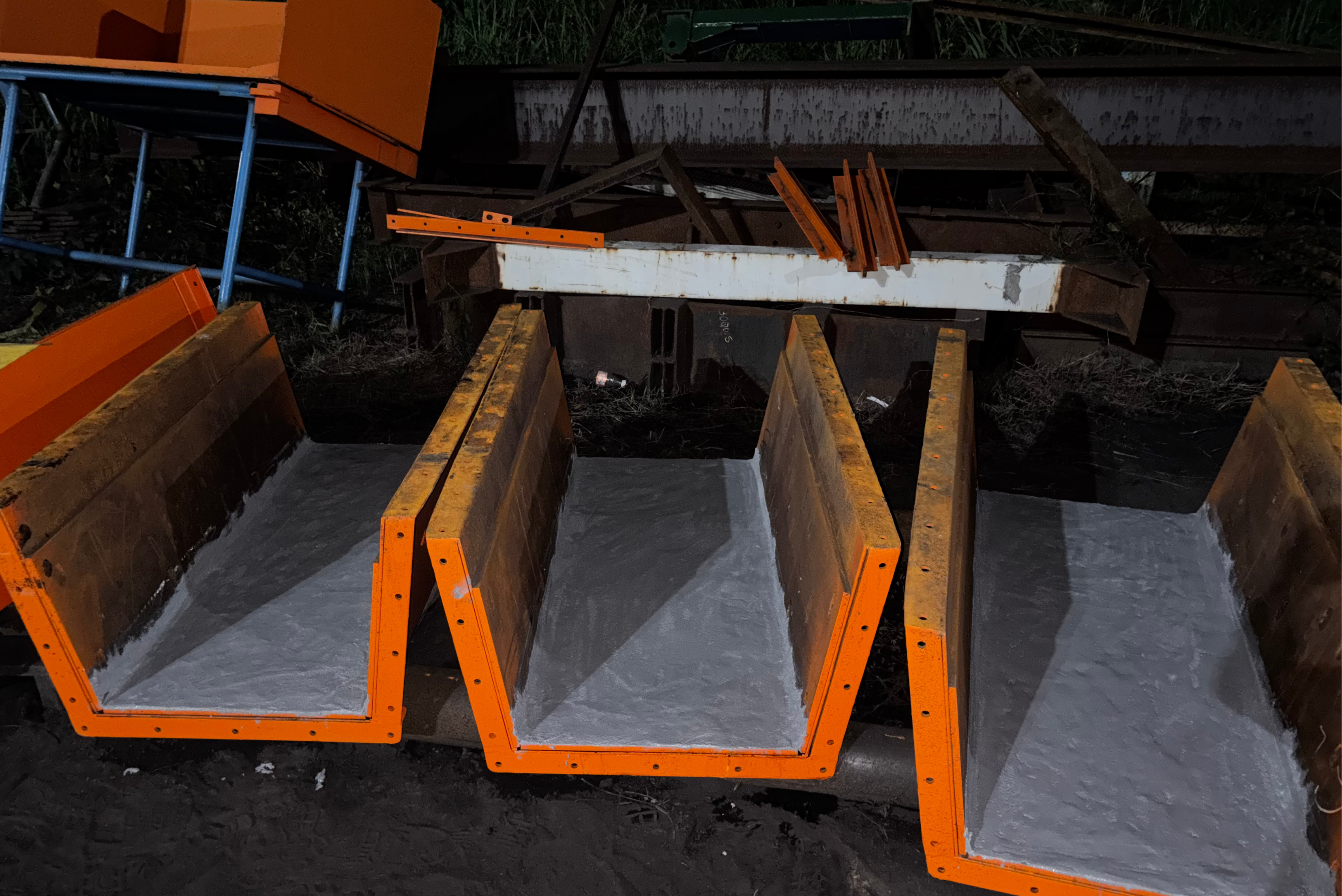 Vários contêineres metálicos de cor laranja com revestimento cinzento no interior dispostos lado a lado sobre uma superfície escura, com equipamentos e estruturas industriais visíveis ao fundo.