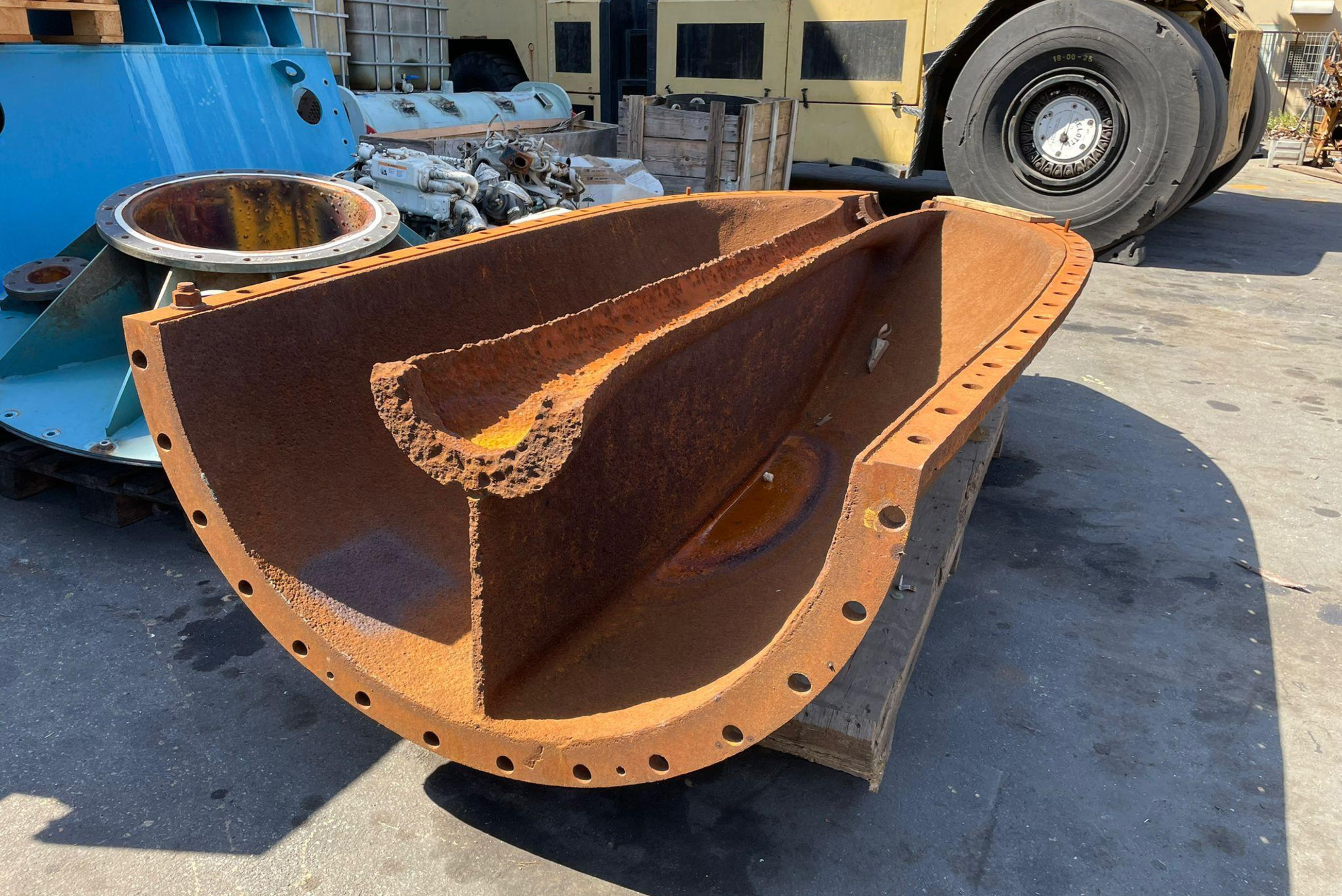 Componente industrial em forma de meia-lua com estrutura metálica avermelhada enferrujada e parafusos de fixação, apoiado em pátio com caminhão azul e equipamentos ao fundo.