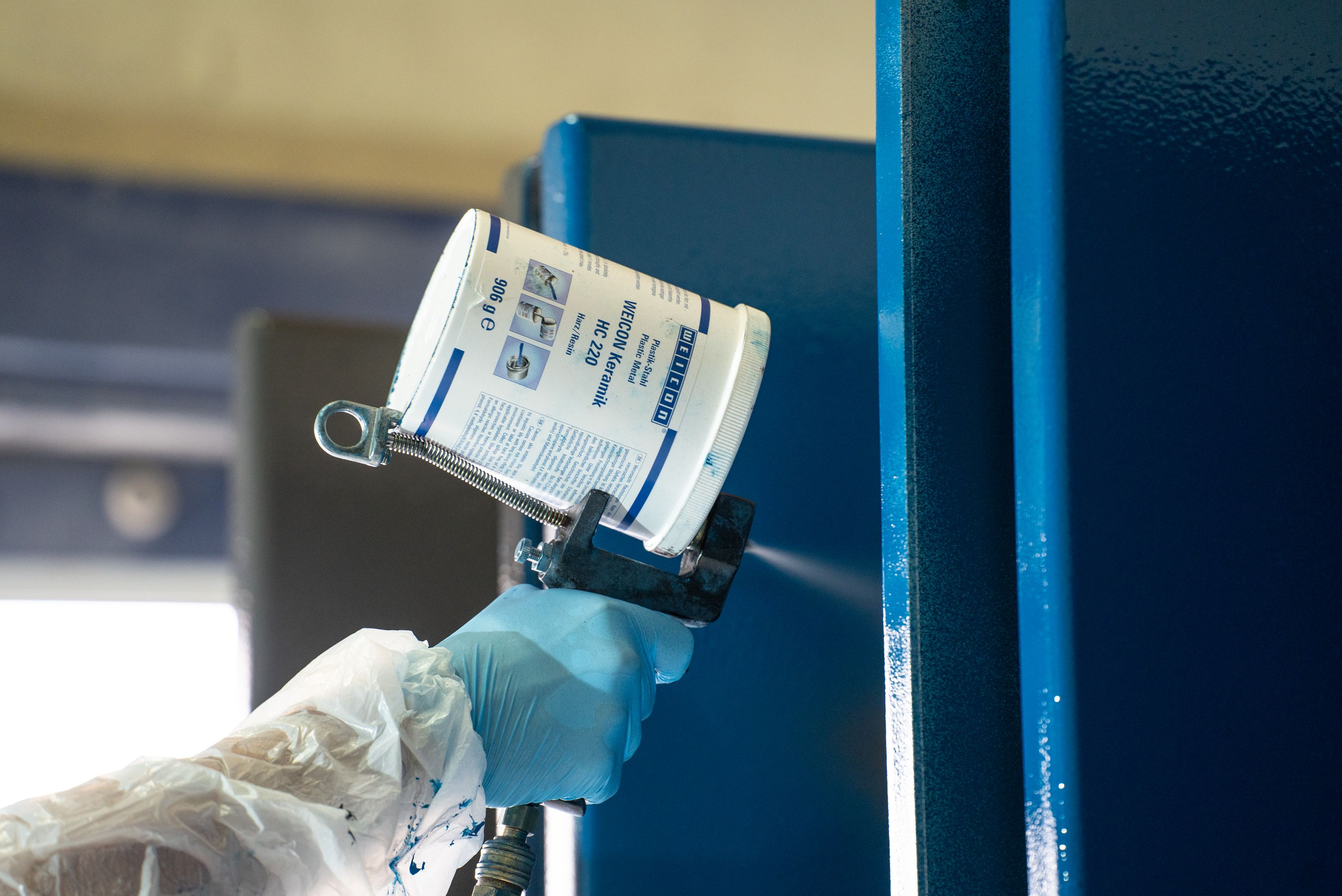 Mão enluvada em branco segurando etiqueta de identificação ou placa de dados presos a estrutura metálica azul em ambiente industrial.