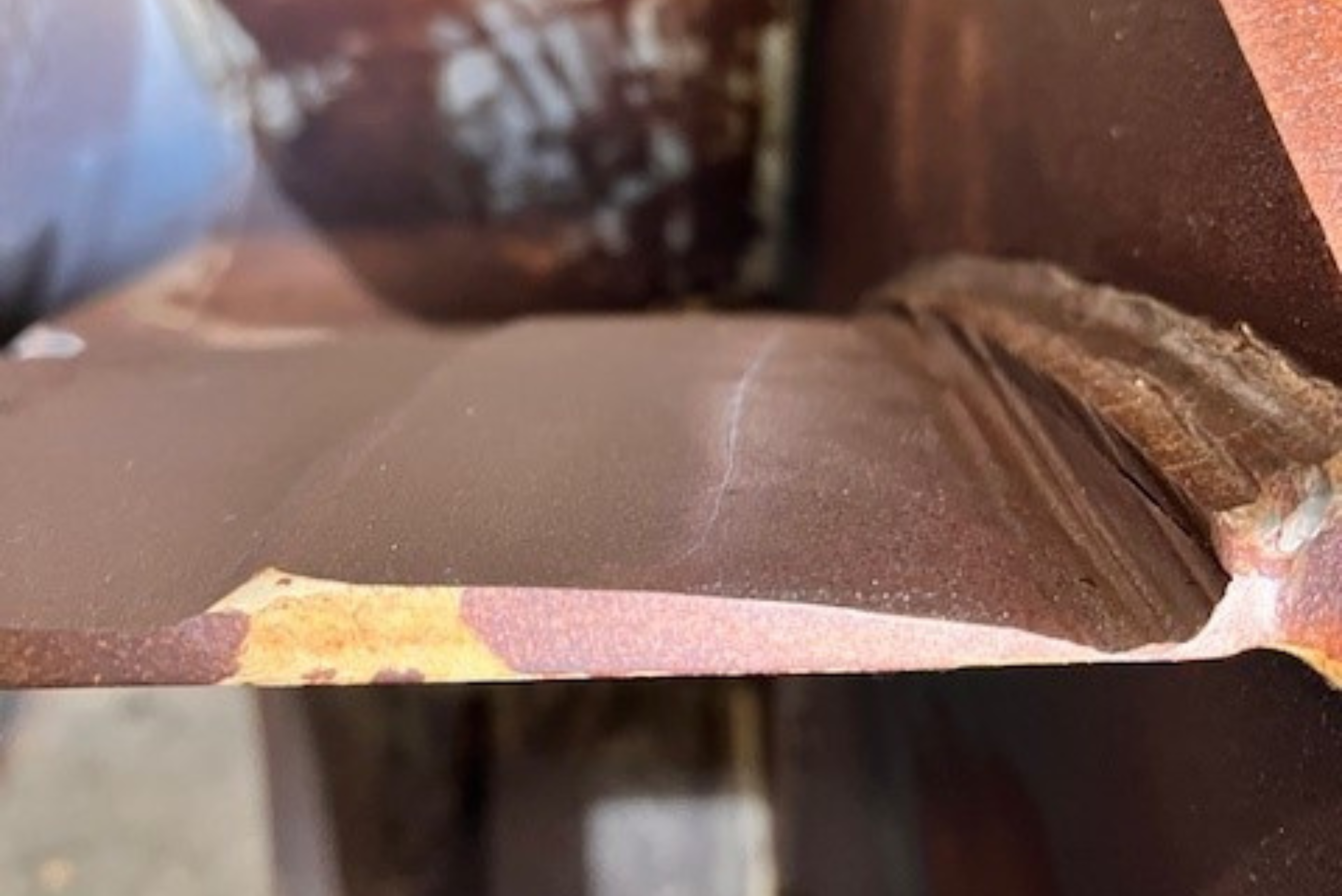 Uma estrutura metálica marrom e robusta, mostrando uma solda grossa e texturizada ao longo da junção com outra seção de metal.