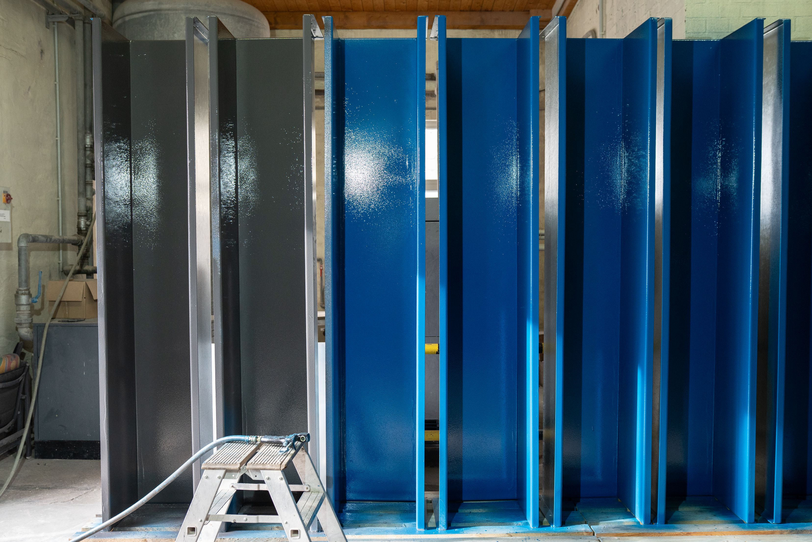 Fileira de painéis metálicos alternando entre cores cinzento escuro e azul brilhante, dispostos verticalmente em ambiente industrial com escada de alumínio e estruturas de suporte visíveis.