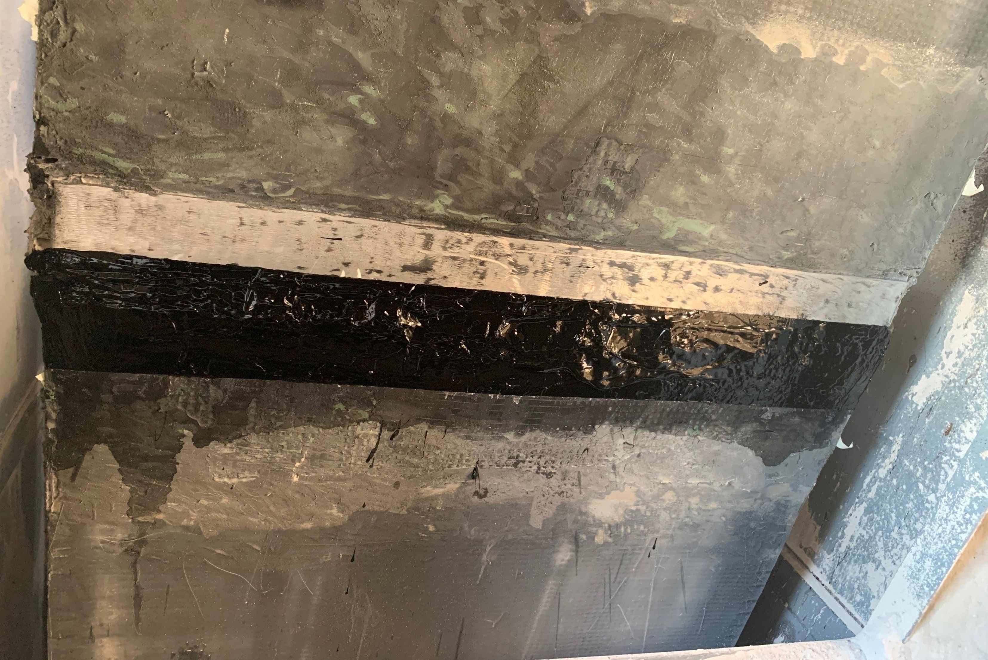 Seção de estrutura metálica com camadas de material cinzento e preto, mostrando corrosão, desgaste superficial e acúmulo de sujeira e resíduos.