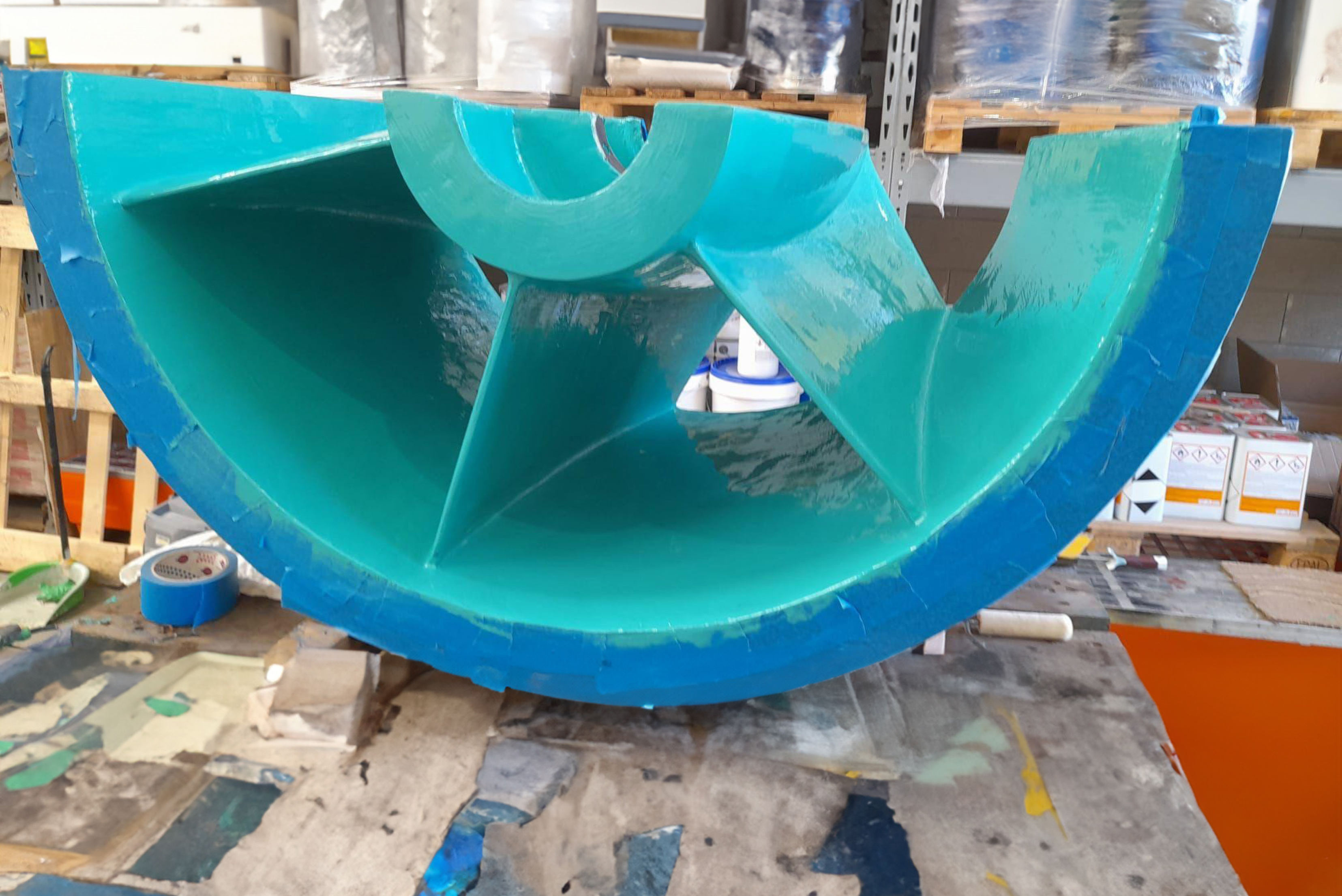 Componente industrial em forma de tigela ou cúpula azul brilhante com pás internas, apoiado em estrutura em ambiente de fábrica com produtos em baldes, ferramentas e equipamentos ao redor.