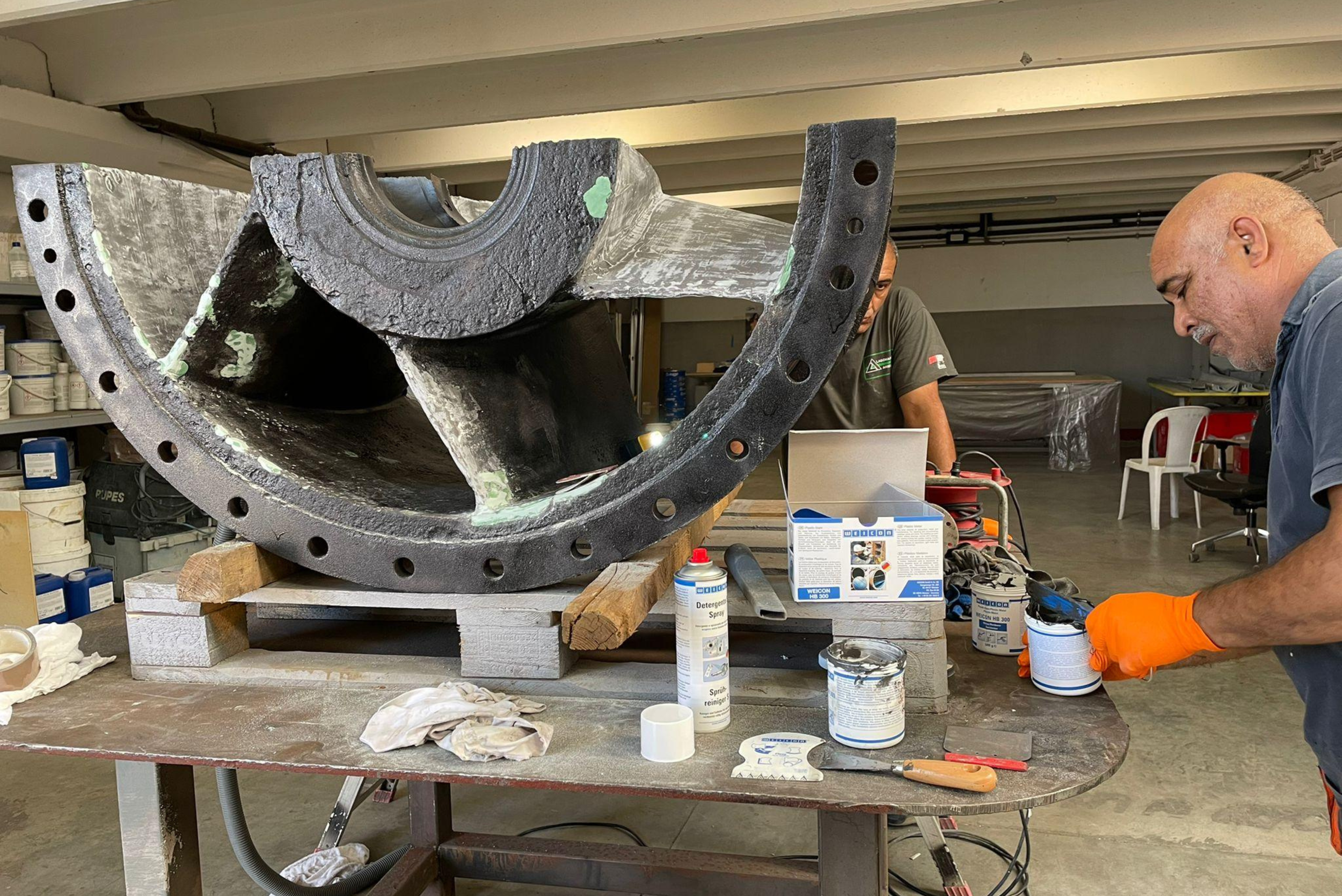 Trabalhador em ambiente de oficina aplicando revestimento ou selante em componente industrial cinzento em forma de arco, com baldes de produto, pincéis e materiais de trabalho dispostos na mesa.