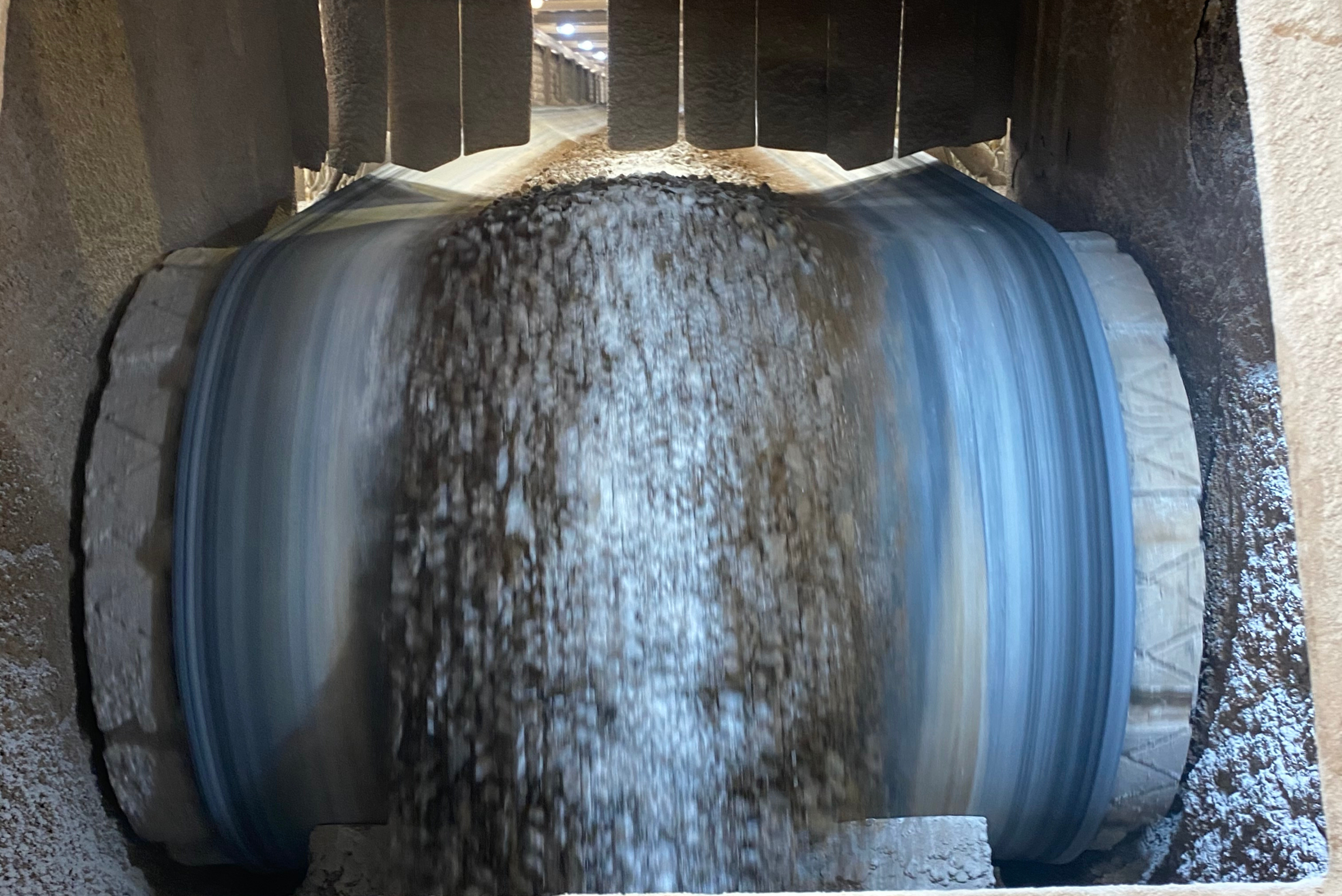 Vista frontal de cilindro industrial cinzento com movimento de material granulado ou pó em seu interior, suspenso por estrutura metálica, mostrando fluxo de material em processo industrial.