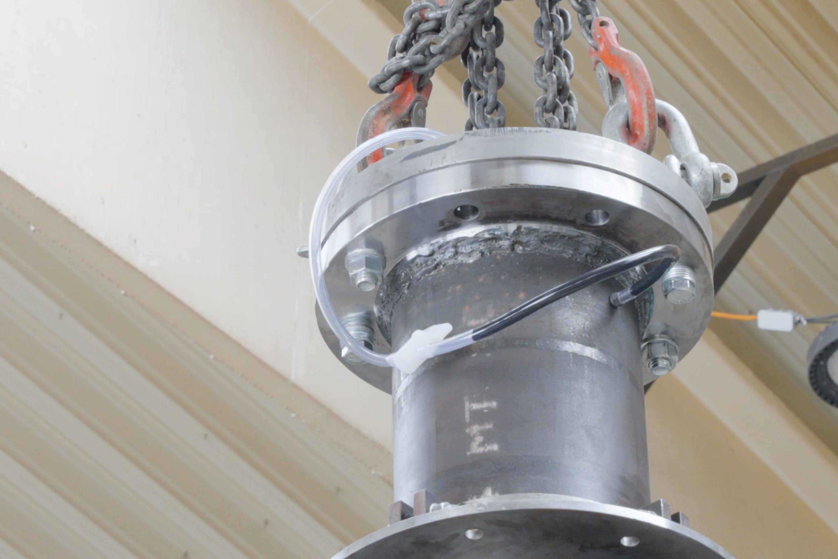 Equipamento cilíndrico cinzento suspenso por correntes metálicas e cabos coloridos presos a uma estrutura branca, instalado no teto com fundo de cobertura ondulada industrial.