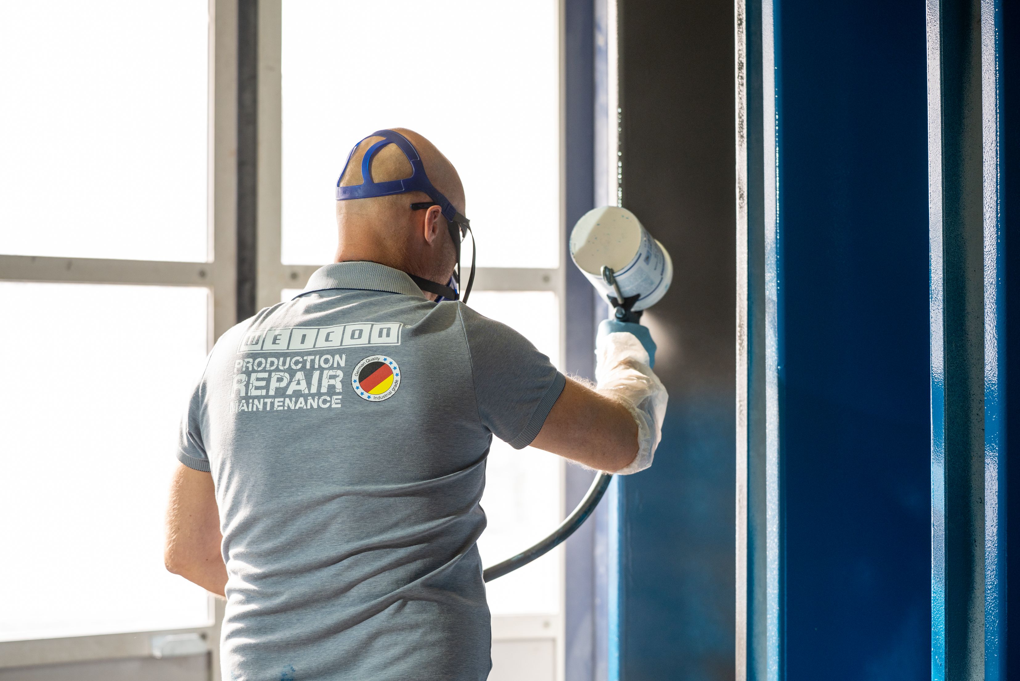 Trabalhador em camiseta cinzenta com texto de reparo, capacete laranja e óculos de proteção realizando inspeção ou manutenção em estrutura metálica azul com ferramenta de medição, próximo a janela com luz natural.