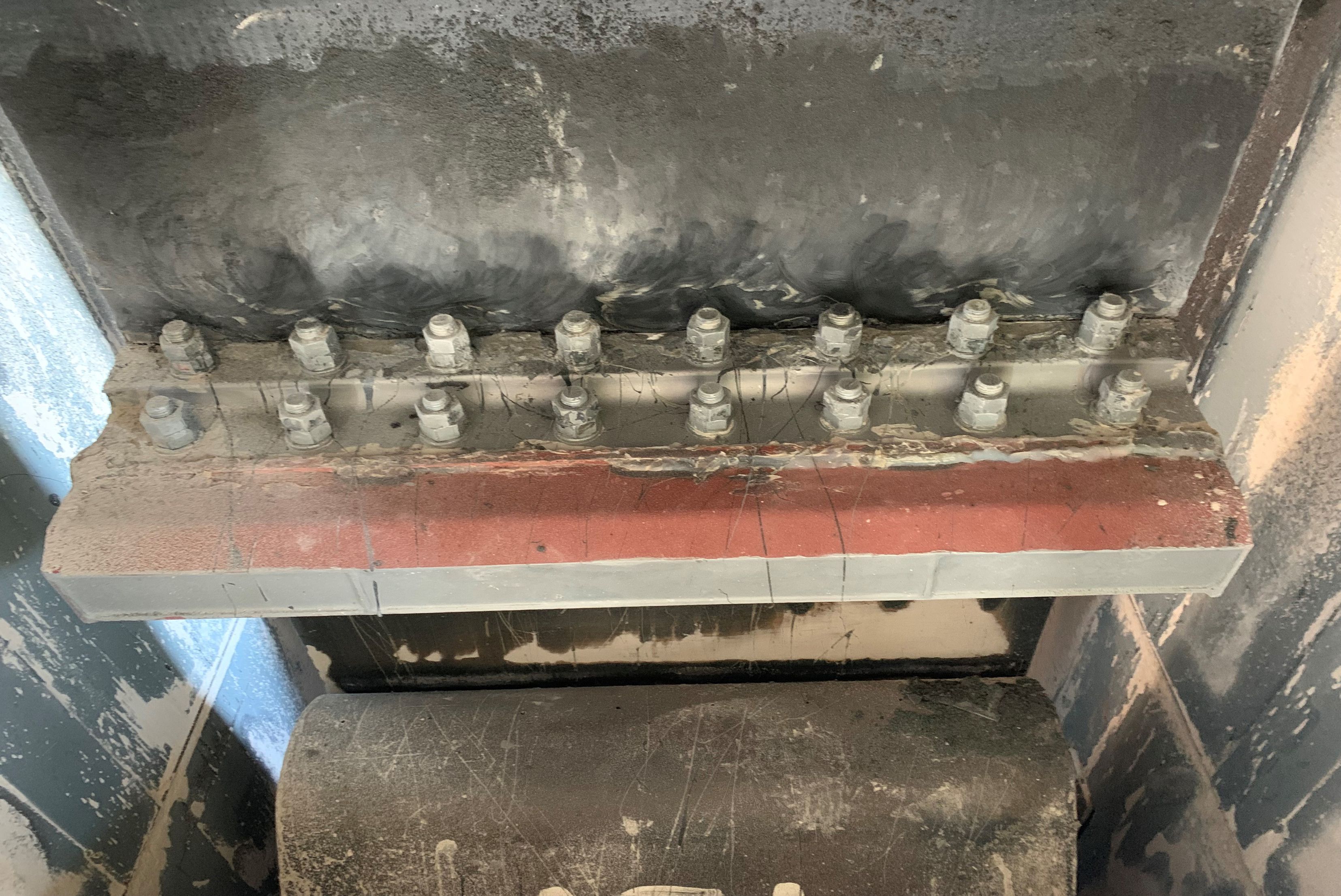 Detalhe de estrutura metálica com fila de parafusos ou rebites metálicos em barra avermelhada, superfícies cinzentas e pretas com corrosão e desgaste, mostrando componentes de montagem industrial