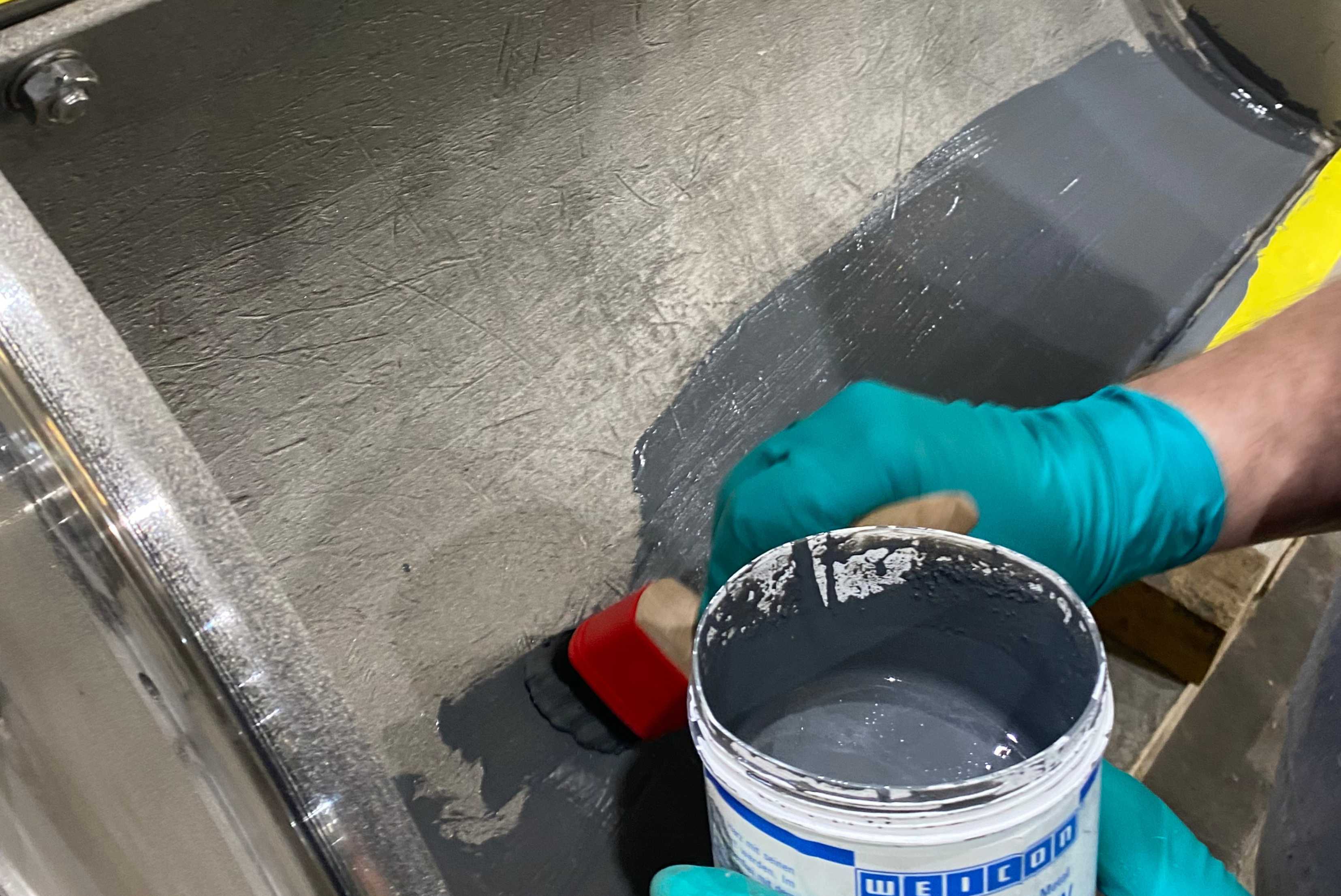 Mão enluvada em azul e vermelho segurando balde branco com produto químico ou revestimento, posicionada sobre superfície metálica cinzenta dentro de contêiner industrial com ferramentas visíveis ao fundo.