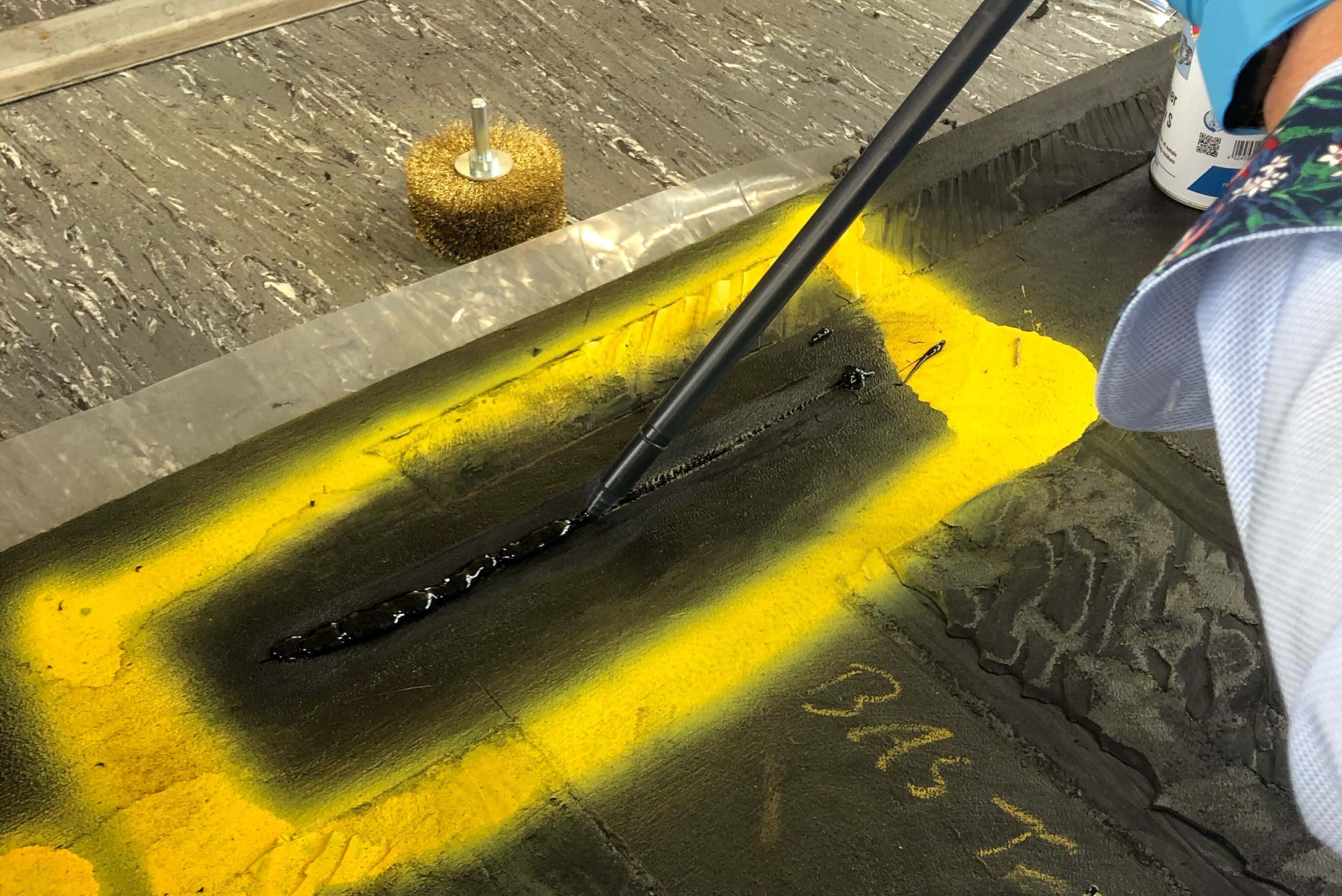 Uma pessoa aplicando um adesivo ou selante escuro em uma fenda dentro de uma área marcada com spray amarelo em uma superfície industrial, com uma escova circular de aço ao fundo