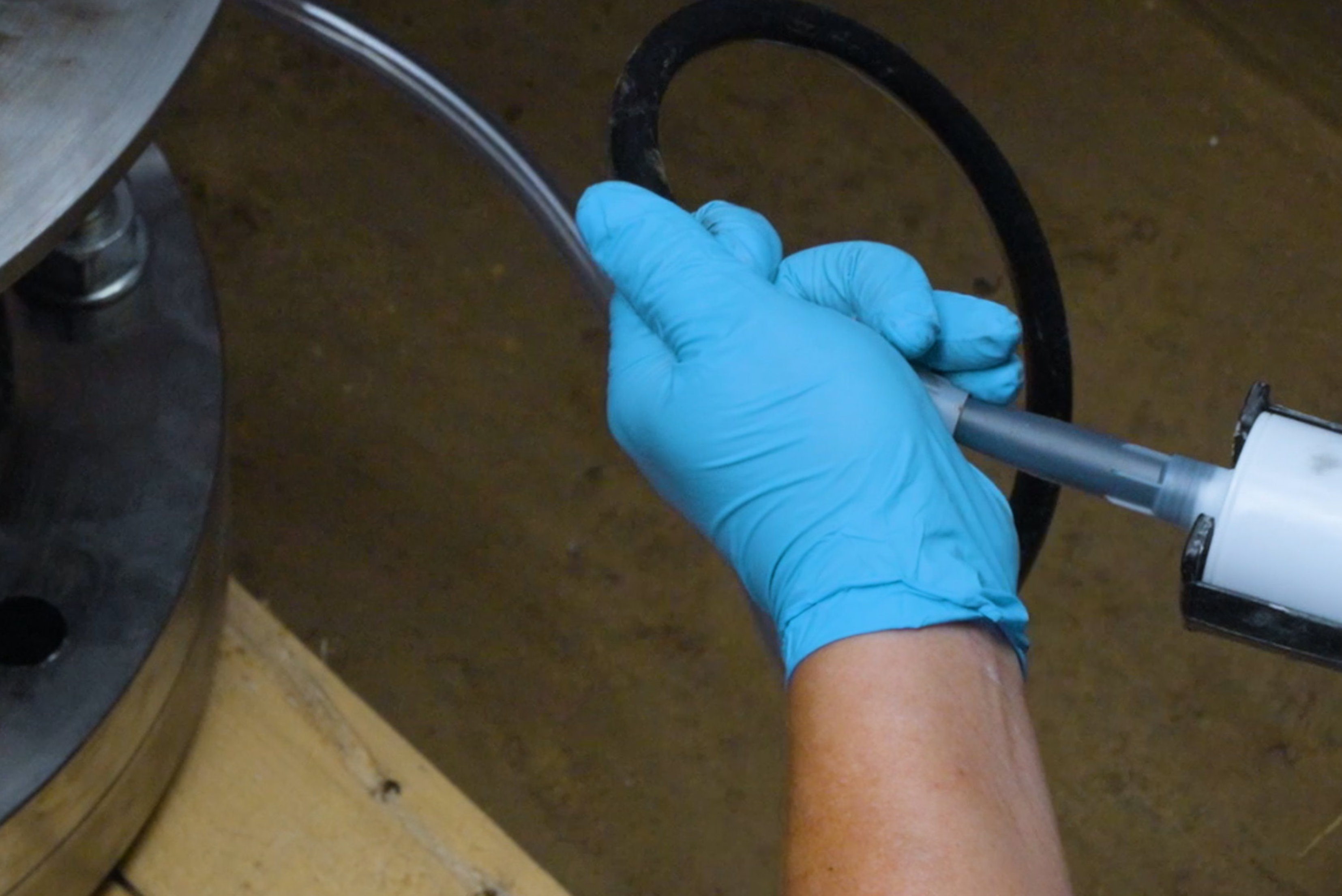 Mão enluvada em azul segurando um tubo ou cabo preto com conector branco, sobre superfície de metal dourado em ambiente industrial.