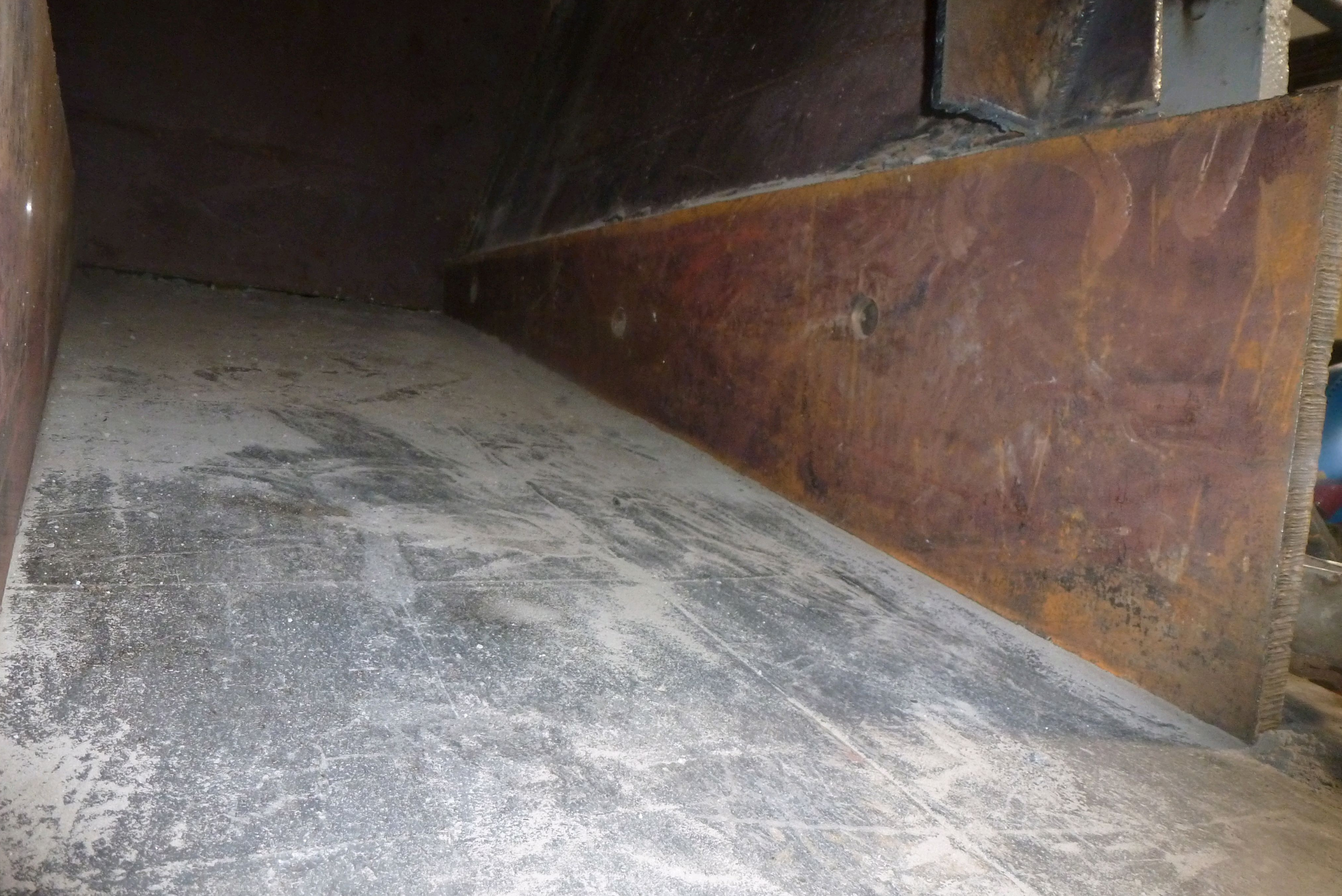 Interior de contêiner metálico com parede lateral enferrujada de cor avermelhada e piso cinzento com marcas de desgaste e sujeira acumulada.