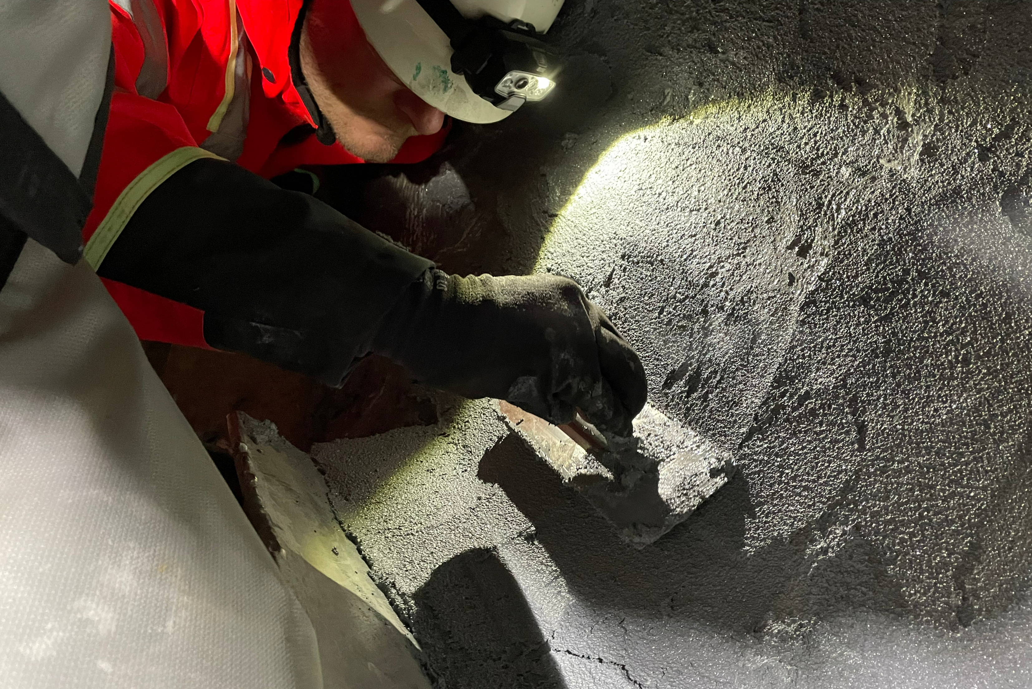 Trabalhador em luvas pretas e vestuário vermelho aplicando ou espalhando revestimento cinzento em pó ou texturizado sobre superfície metálica em ambiente industrial.
