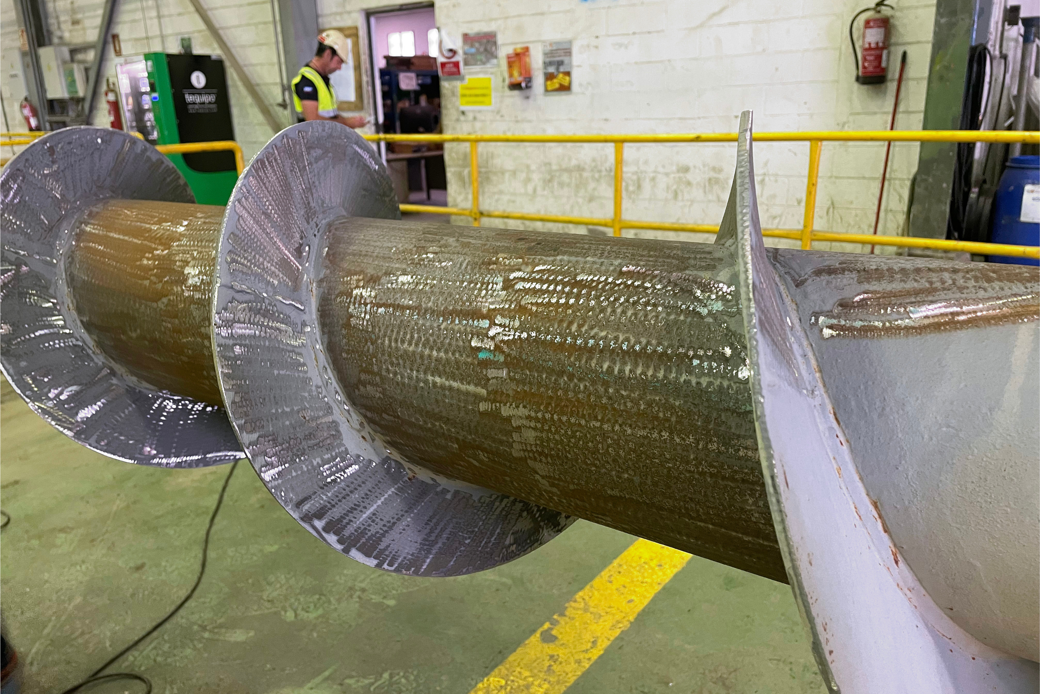 Cilindro industrial metálico envolto em plástico protetor transparente, com flanges circulares nas extremidades, apoiado em estrutura de suporte em piso industrial verde com demarcação amarela.
