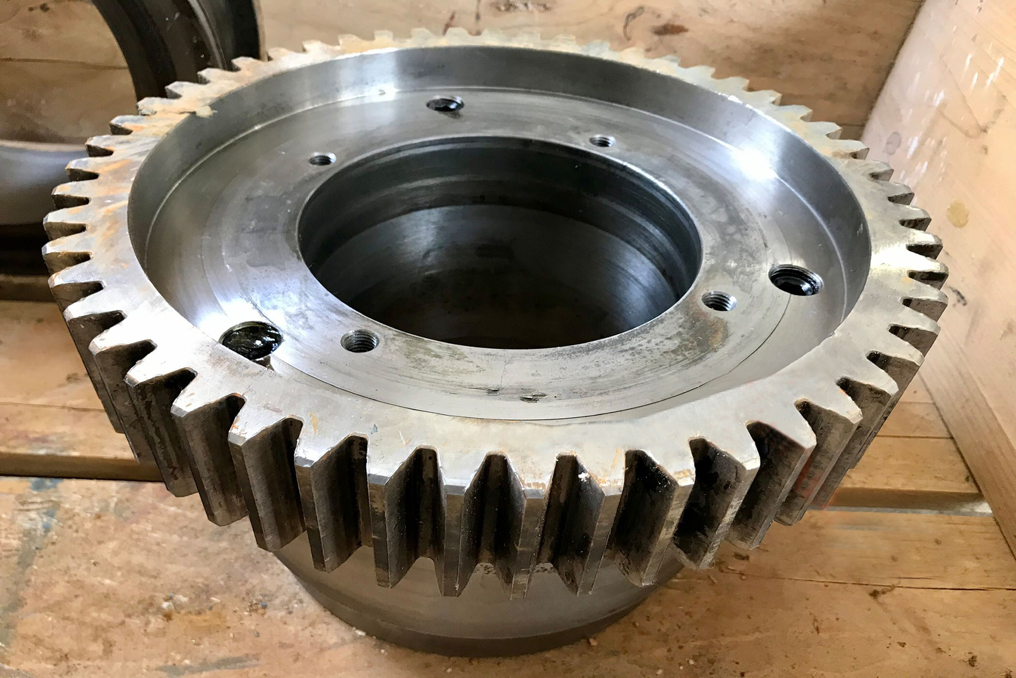 Engrenagem industrial metálica prateada com dentes grandes radiais, abertura central circular, apoiada em superfície de madeira em ambiente de oficina ou armazém.
