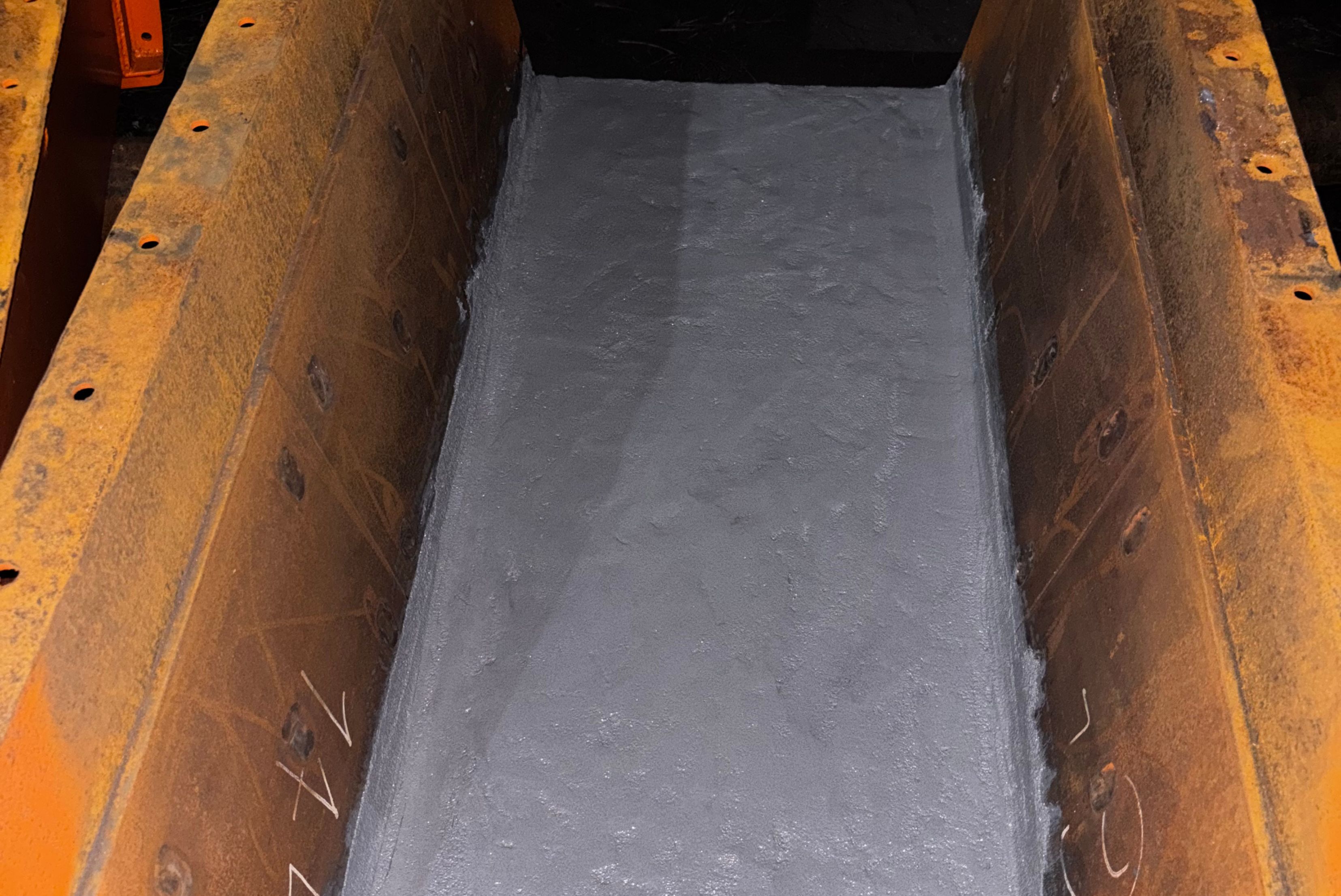 Vista superior de um contêiner de metal oxidado com paredes avermelhadas, mostrando o interior vazio com piso de metal cinzento liso e marcas de desgaste nas laterais enferrujadas.