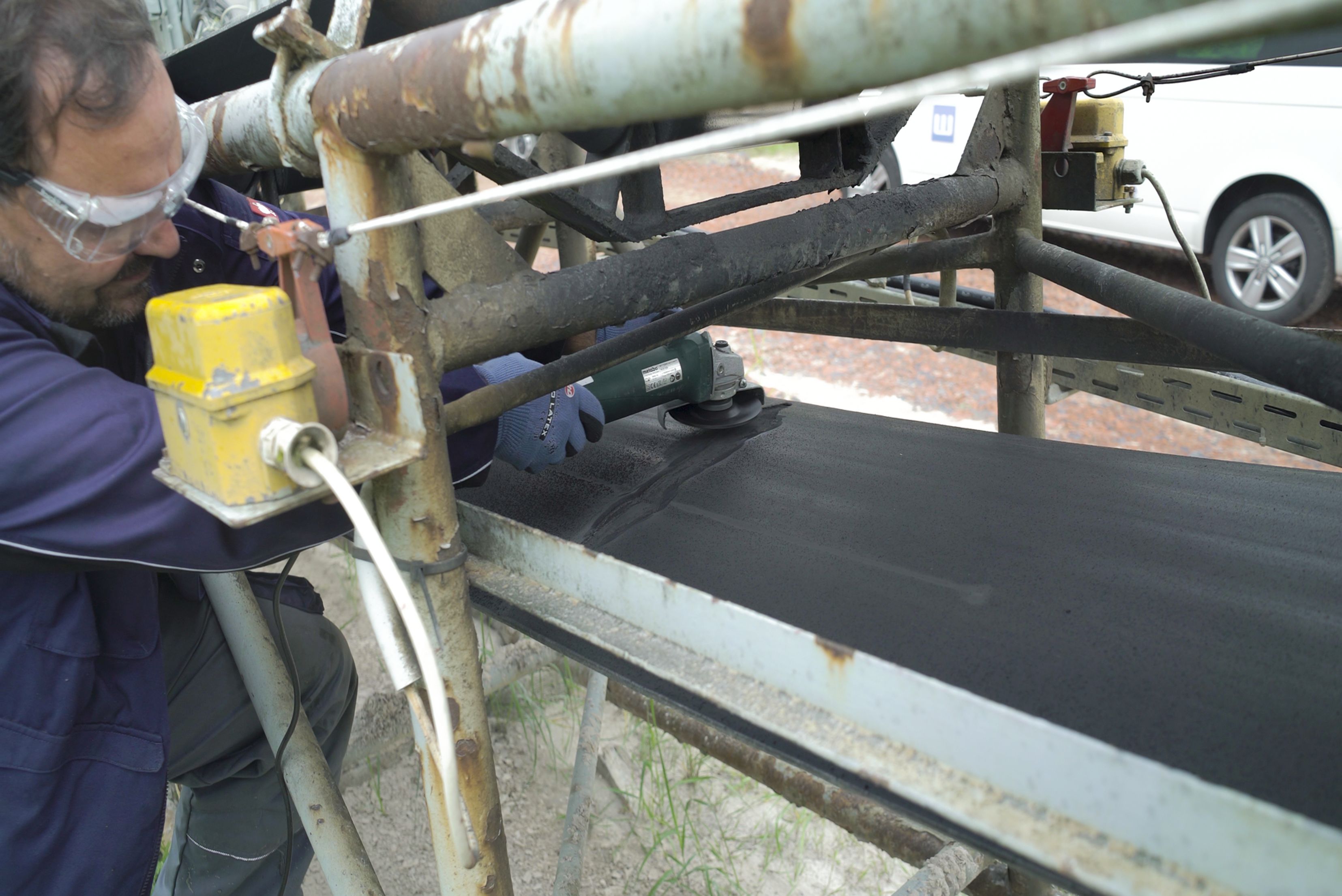 Trabalhador em uniforme azul e colete amarelo operando ou inspecionando equipamento industrial metálico com tubo enferrujado, em ambiente externo com veículo branco visível ao fundo.