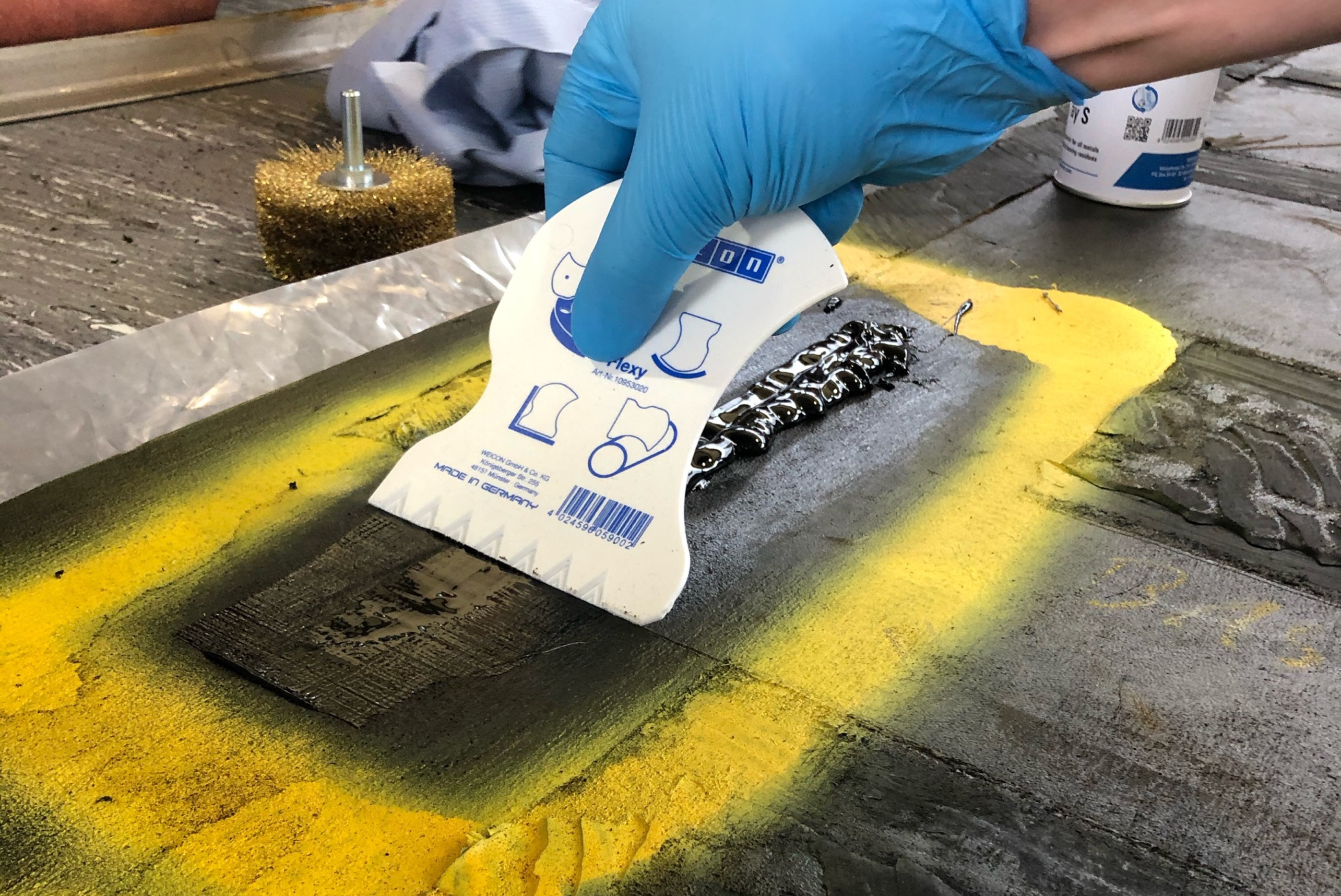 Eine mit blauen Handschuhen bekleidete Hand benutzt einen weißen Spachtel mit blauer Aufschrift, um eine dunkle Masse auf einer mit gelber Farbe markierten Industriefläche glatt zu streichen.