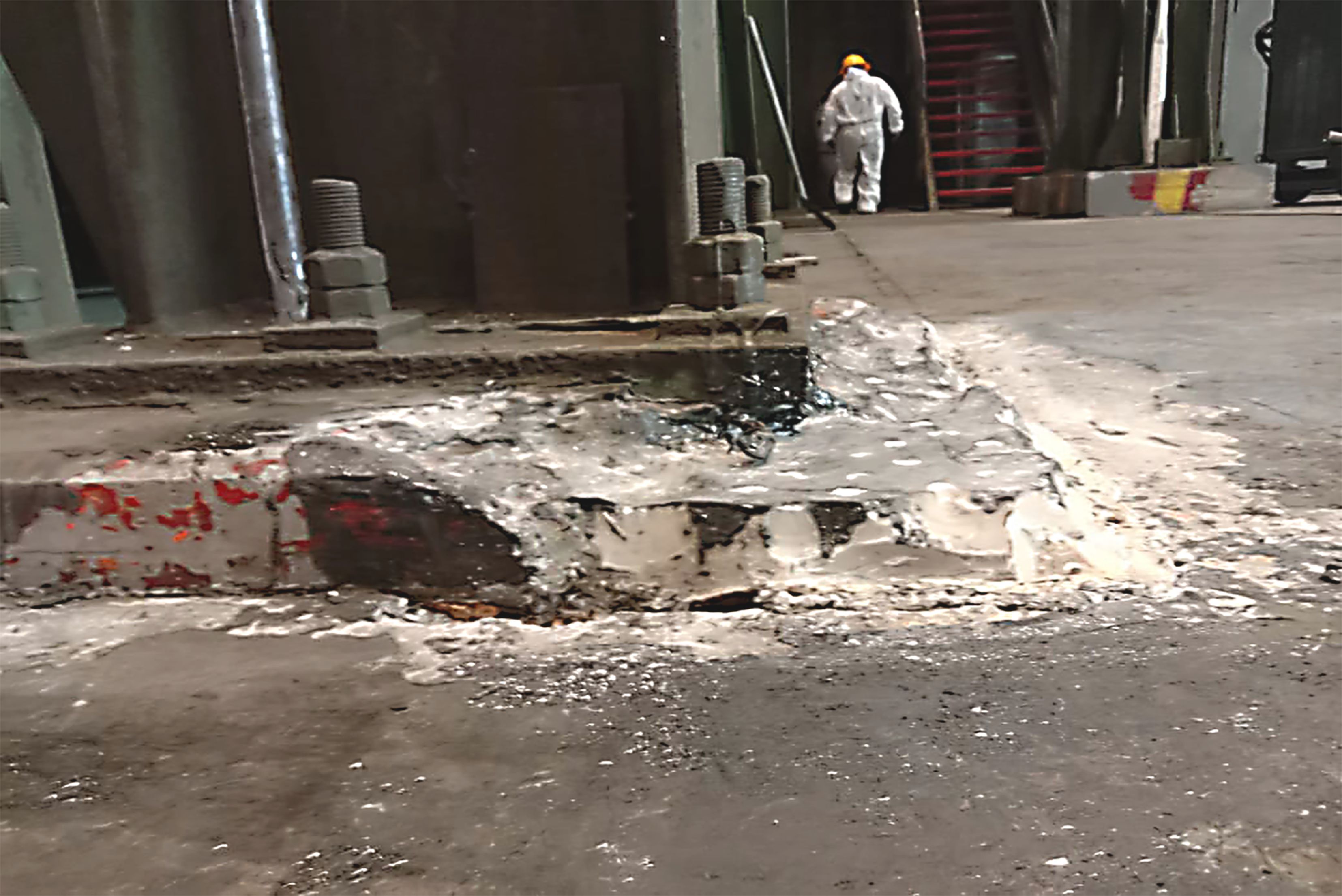 Vista de pátio industrial com buraco ou escavação no piso de concreto, material solto espalhado, estruturas metálicas enferrujadas ao fundo, trabalhador ao longe e edifício industrial.