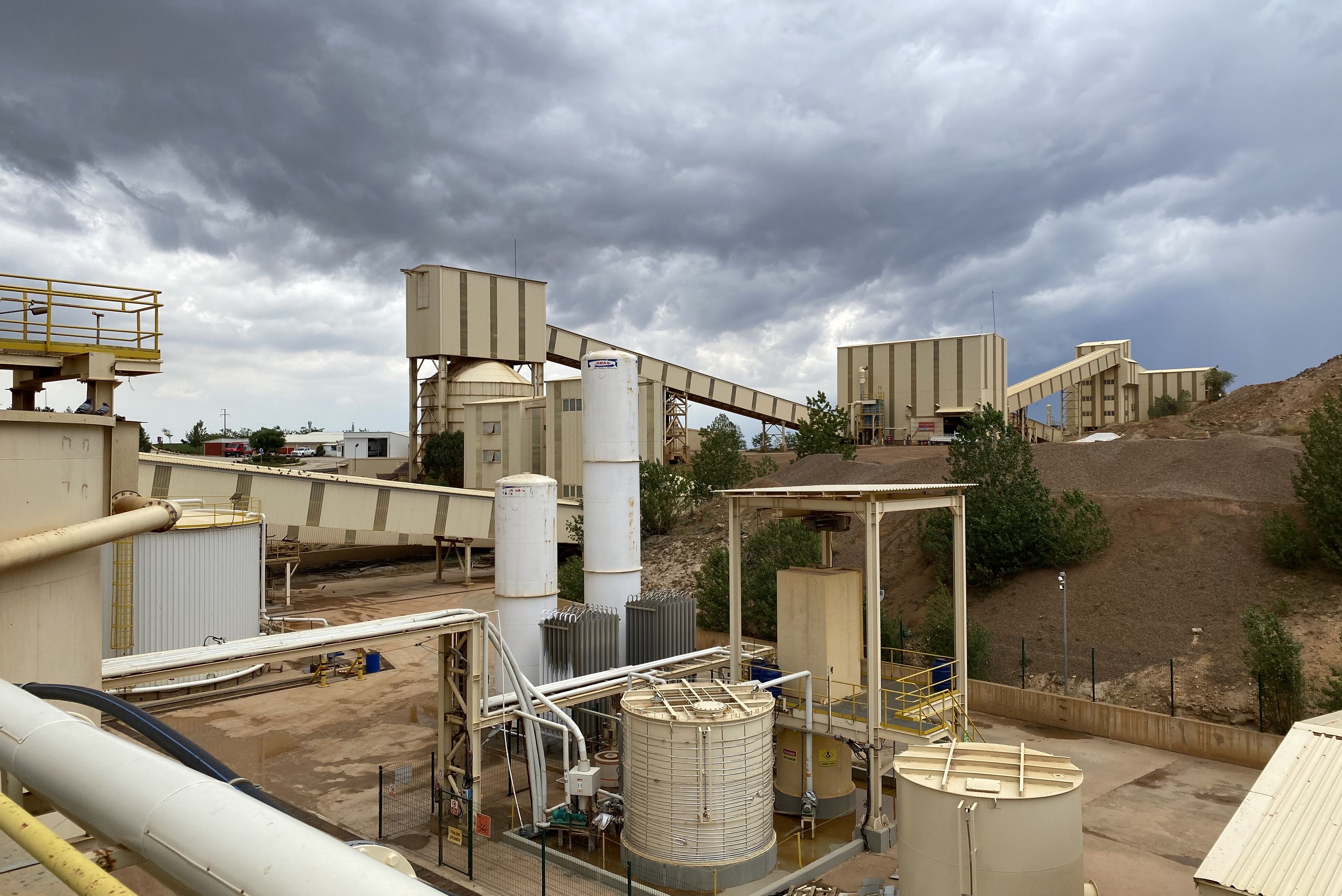 Vista panorâmica de instalação industrial com múltiplos silos e equipamentos de processamento em tons bege e cinzento, estruturas de metal, céu nublado ao fundo e terreno argiloso ao redor.