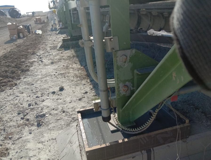 Uma estrutura industrial verde de suporte de metal ao ar livre, ancorada em uma base de concreto com uma moldura de madeira, situada ao lado de um caminho de terra em um canteiro de obras ou área industrial.