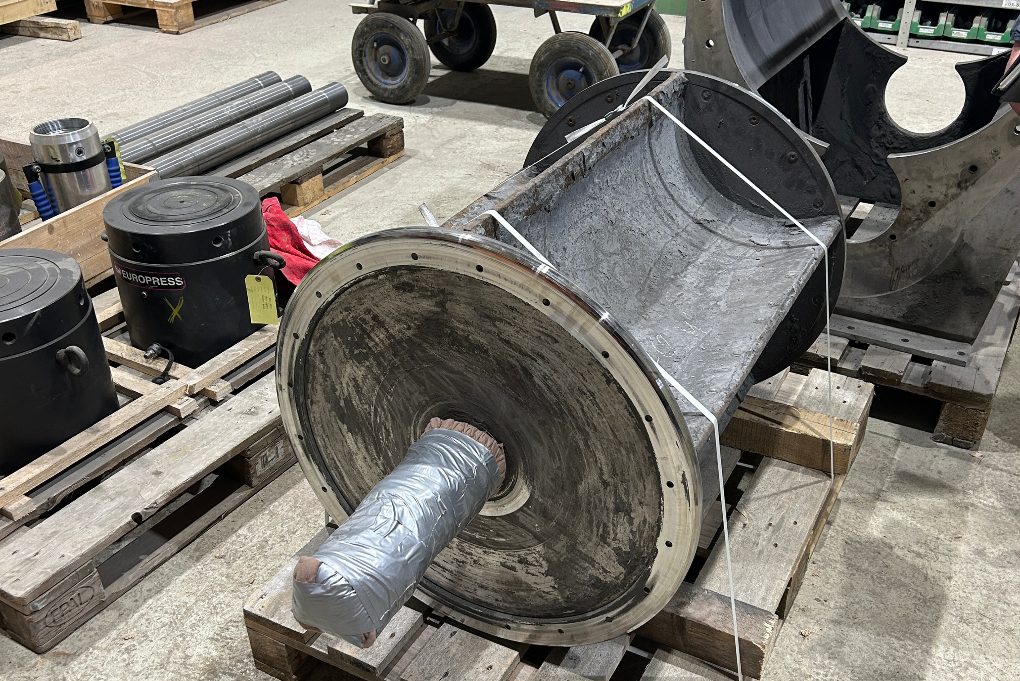 Componente industrial desmontado mostrando cilindro metálico cinzento com eixo central, flanges de metal, superfícies com desgaste e corrosão, junto a outras peças e equipamentos em pátio industrial.
