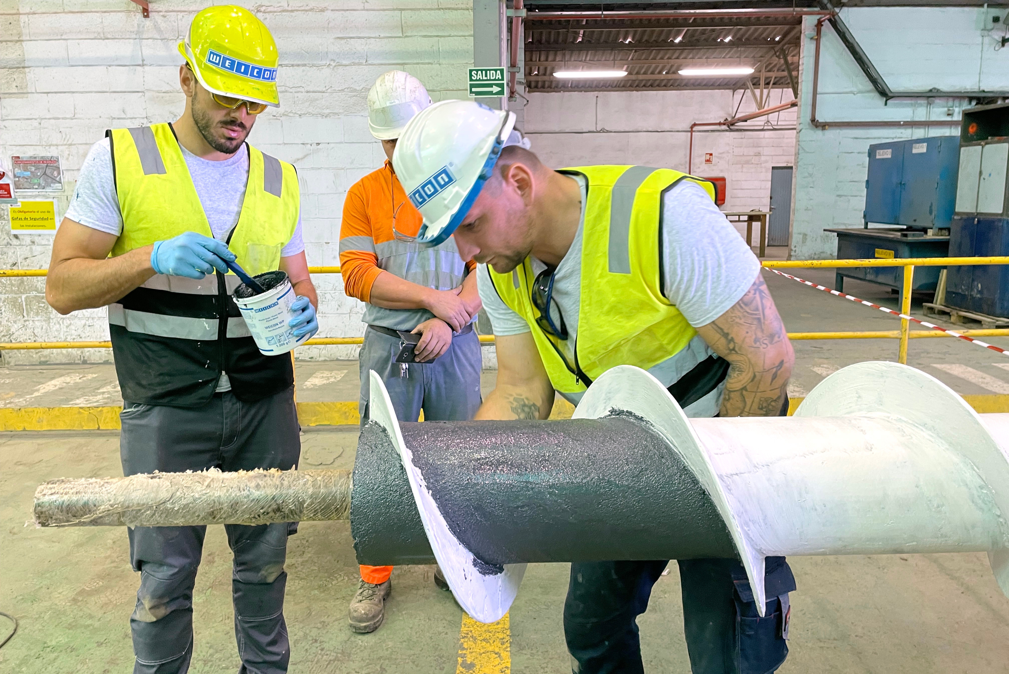 Três trabalhadores em equipamento de proteção amarelo fluorescente e capacetes inspecionando cilindro industrial metálico com revestimento cinzento e branco, em ambiente de fábrica com demarcação de segurança amarela.